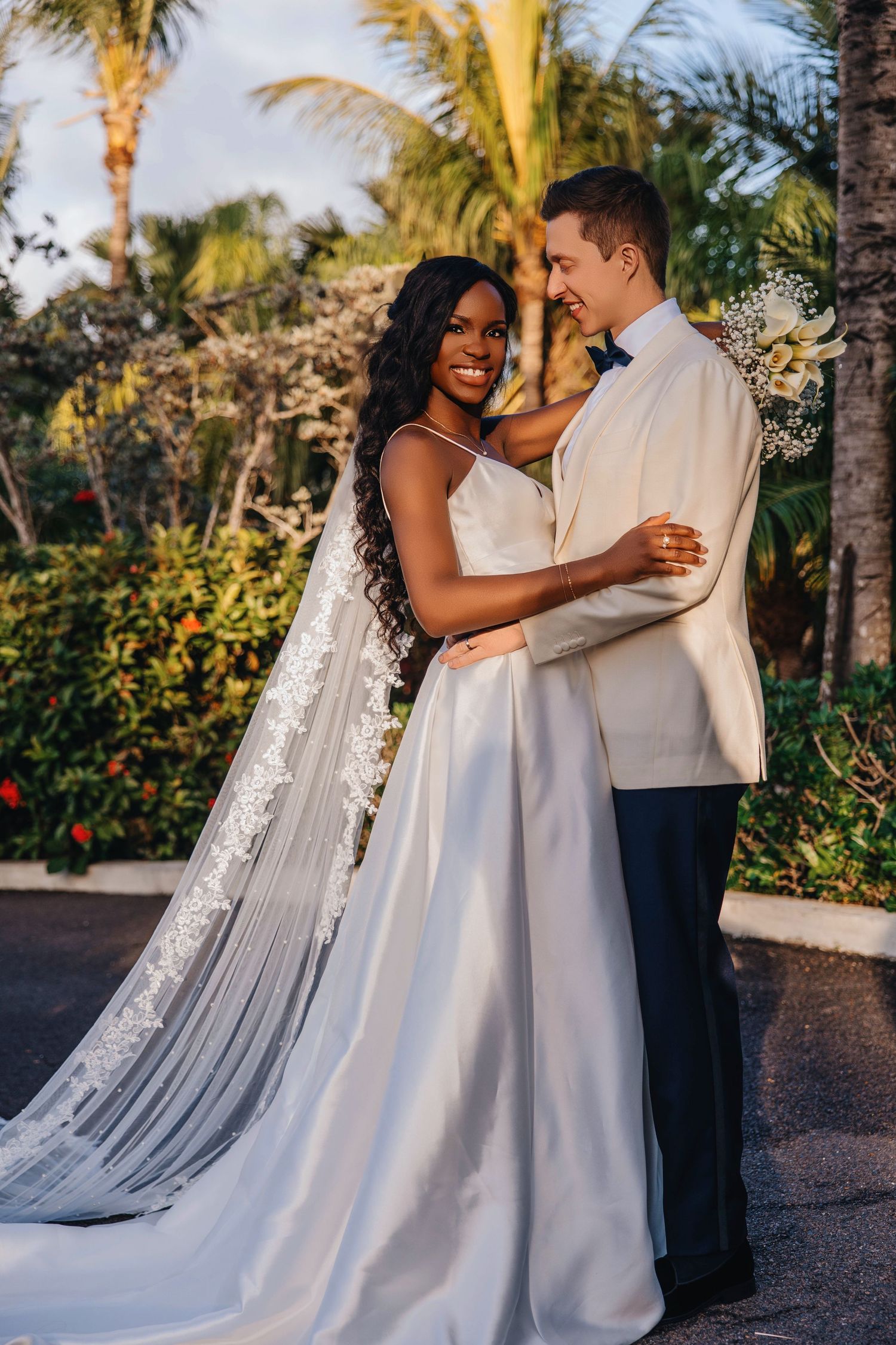 Bride in flowing white gown and groom in white jacket share romantic moment among palm trees.
