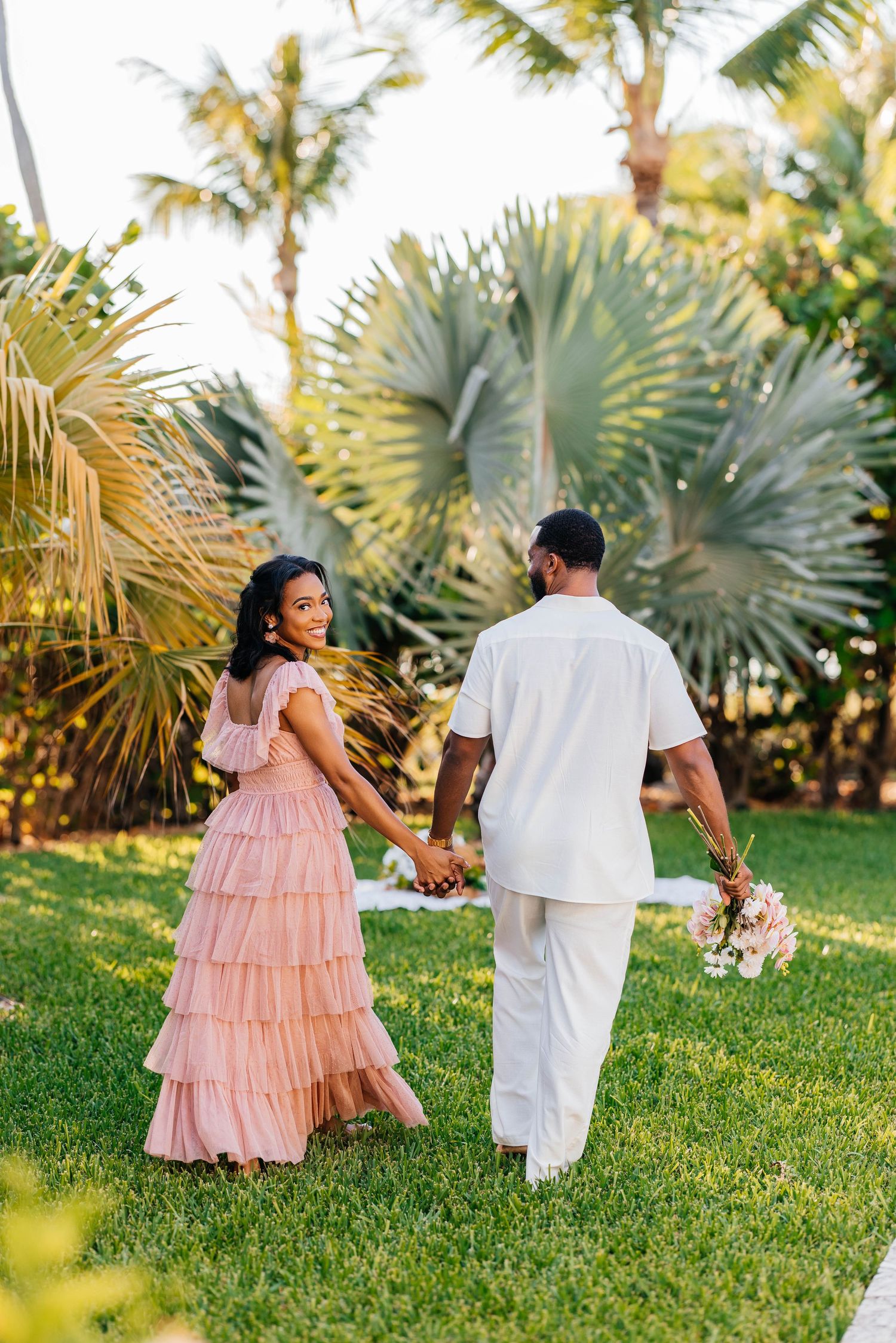 A couple holding hands walks on grass among tropical palm trees and greenery in romantic garden setting.