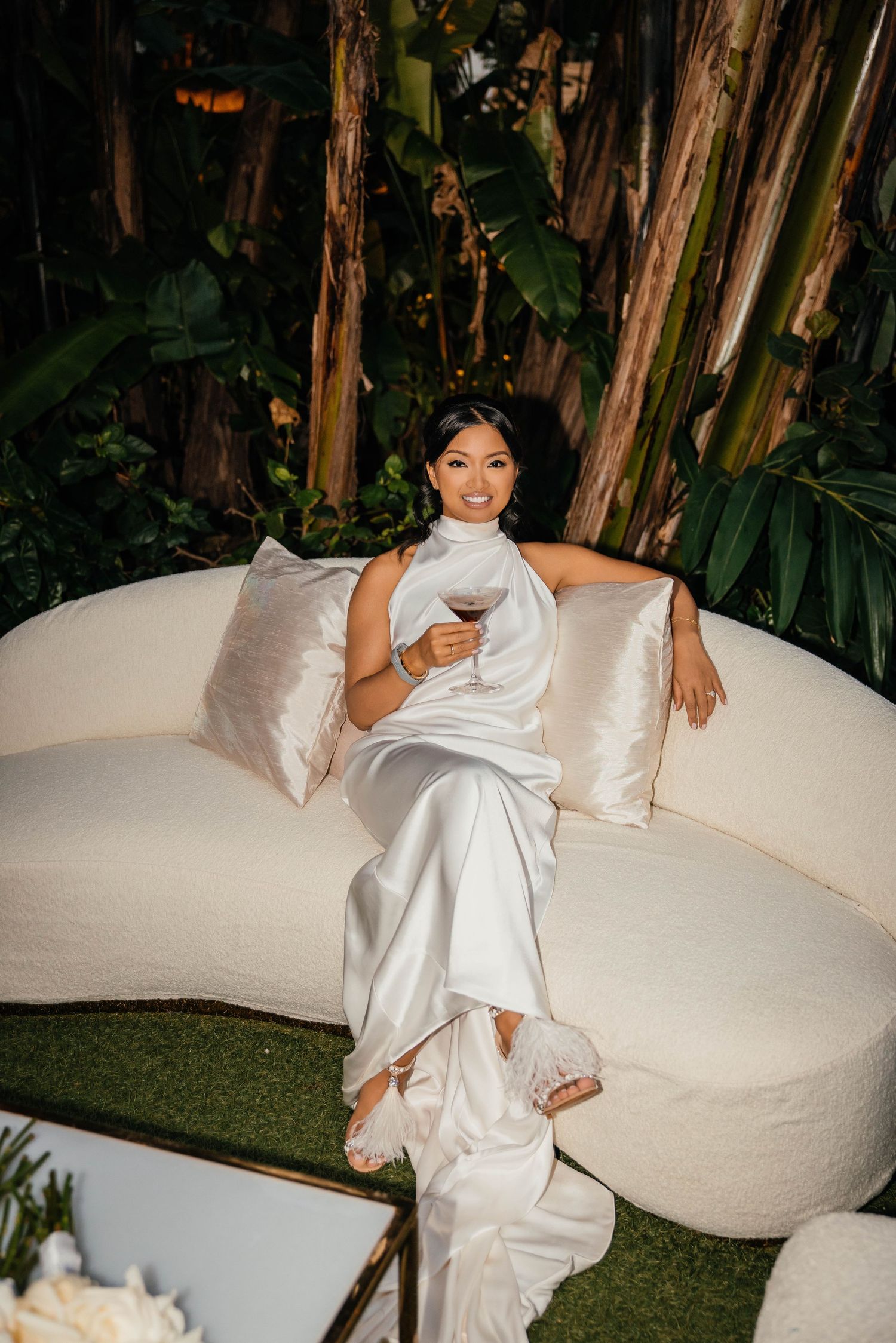 Elegant woman in white satin gown holding wine glass, relaxing on cream curved sofa surrounded by tropical greenery.