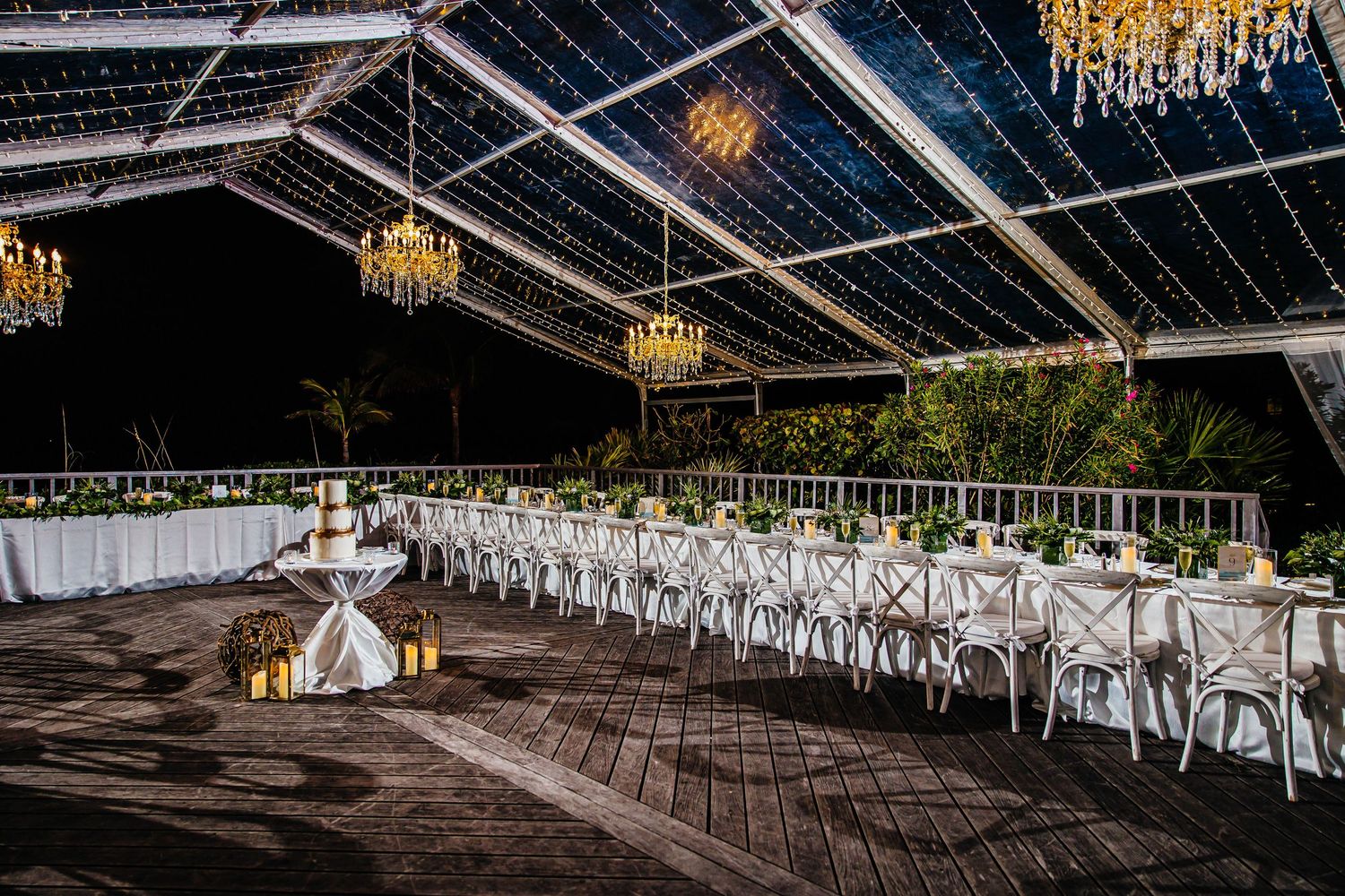 Elegant outdoor wedding reception under a clear tent with chandeliers, string lights, and long white tables on a wooden deck.
