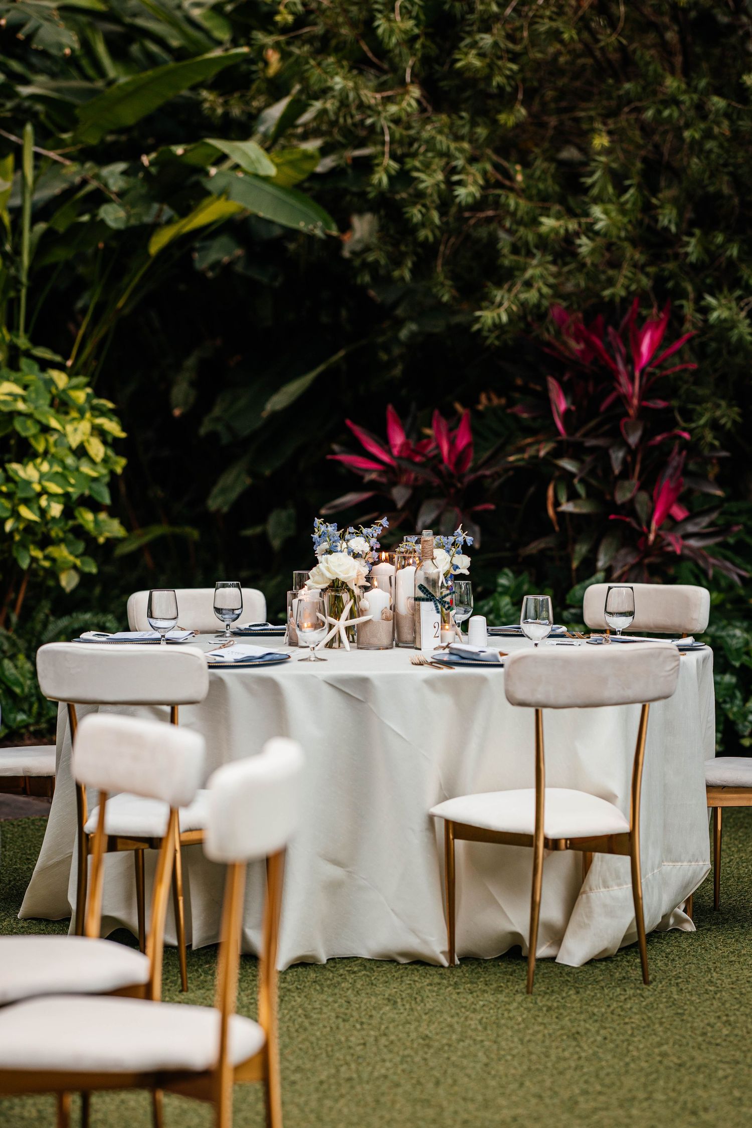 Elegant outdoor wedding reception table with white linen, gold chairs, floral centerpiece, and lush tropical greenery backdrop.