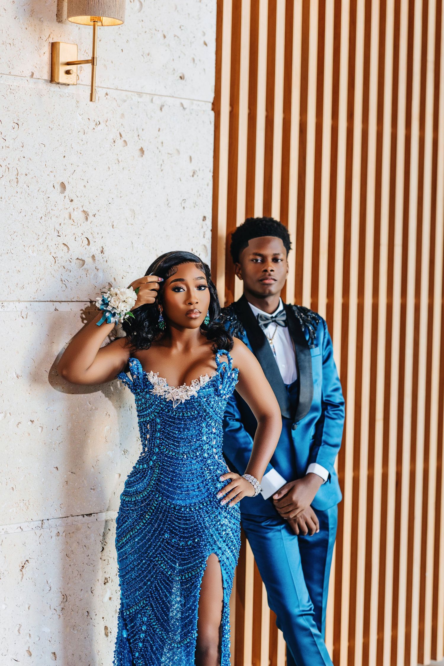 Elegant couple in coordinating blue formal attire poses against wooden slat wall with white stone accents.