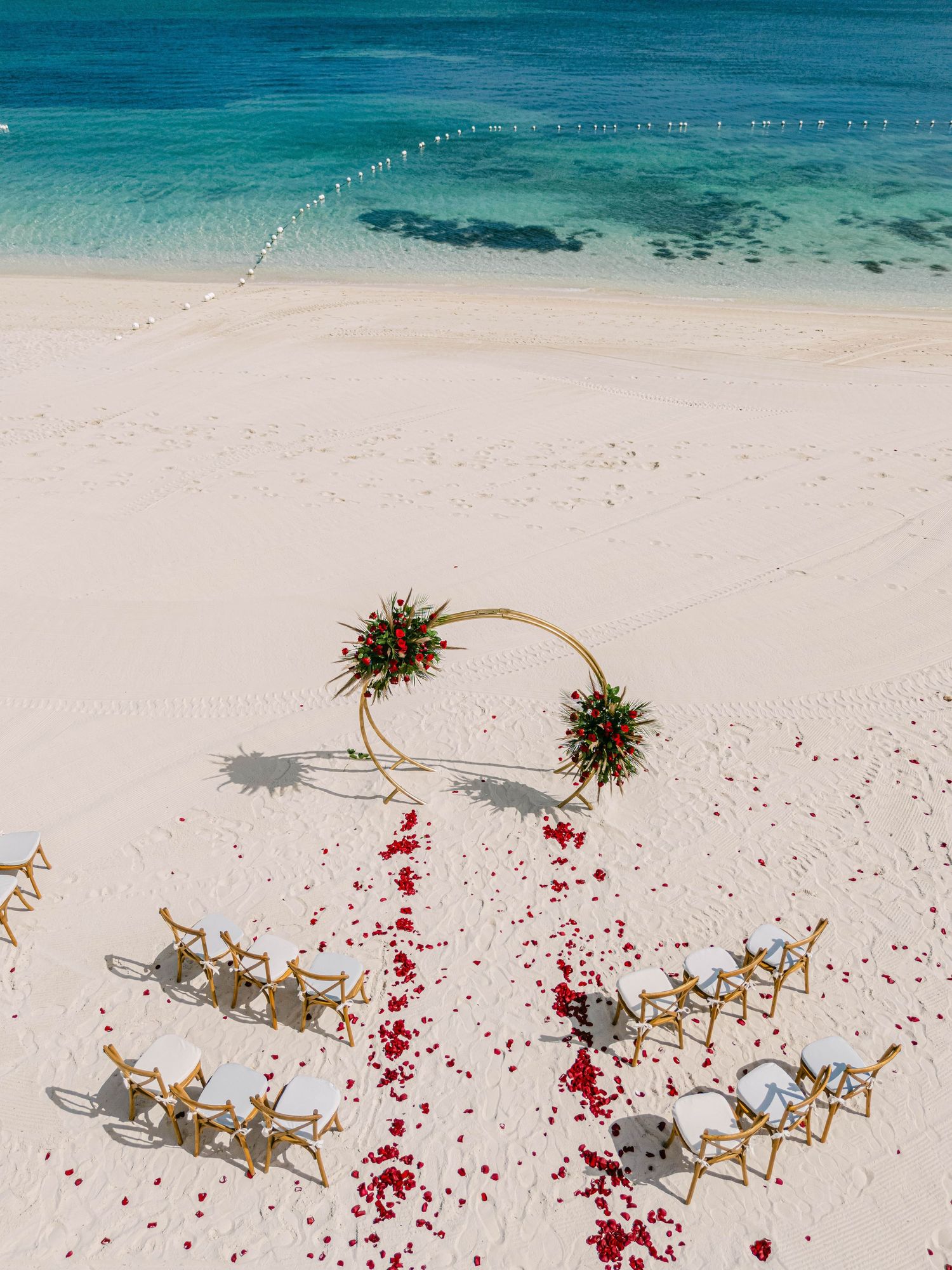 Aerial view of a beach wedding setup with a gold circular arch, red rose petal aisle, and wooden chairs on white sand.