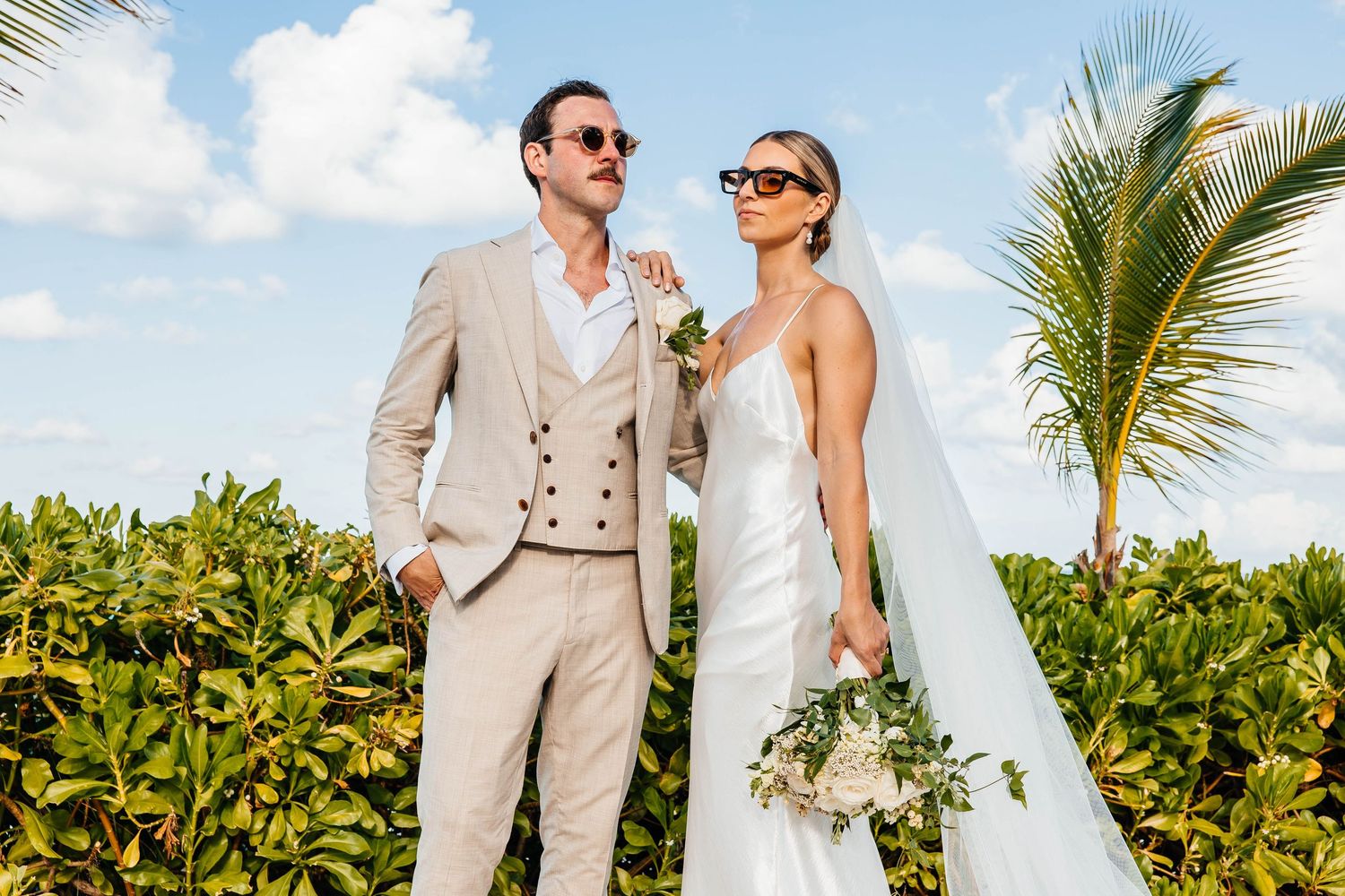 Destination wedding couple in beige and white attire with palm trees and blue sky.