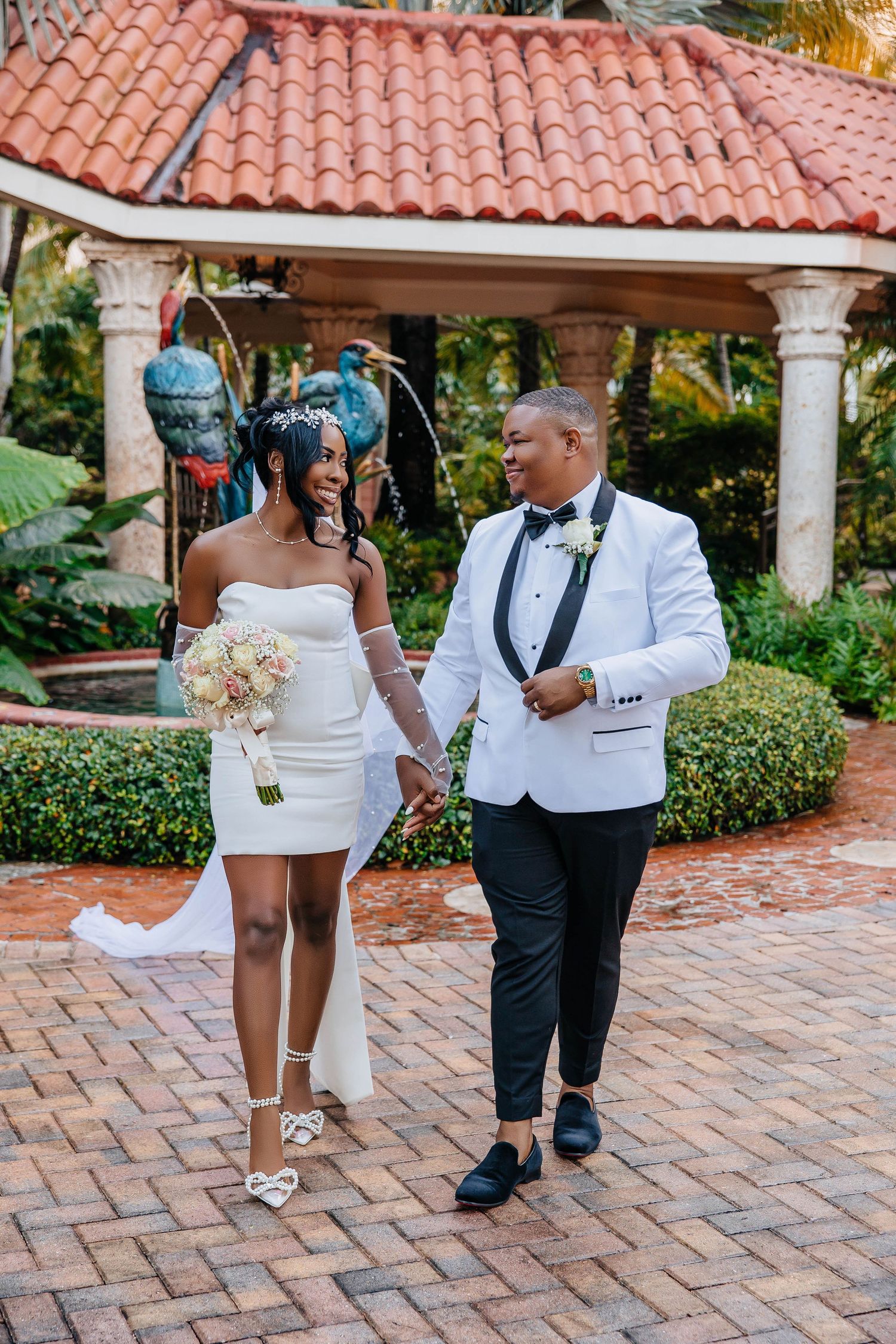 A newly married couple walking together on brick pavers under a Spanish-style pavilion.