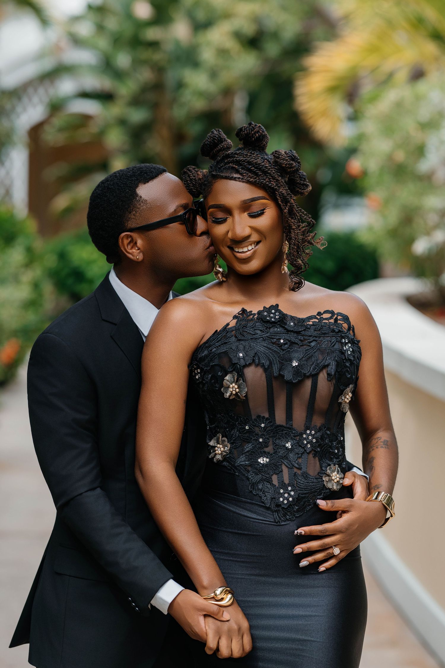 Elegant couple in black formal attire sharing a romantic moment outdoors, man kissing woman's cheek as she smiles.