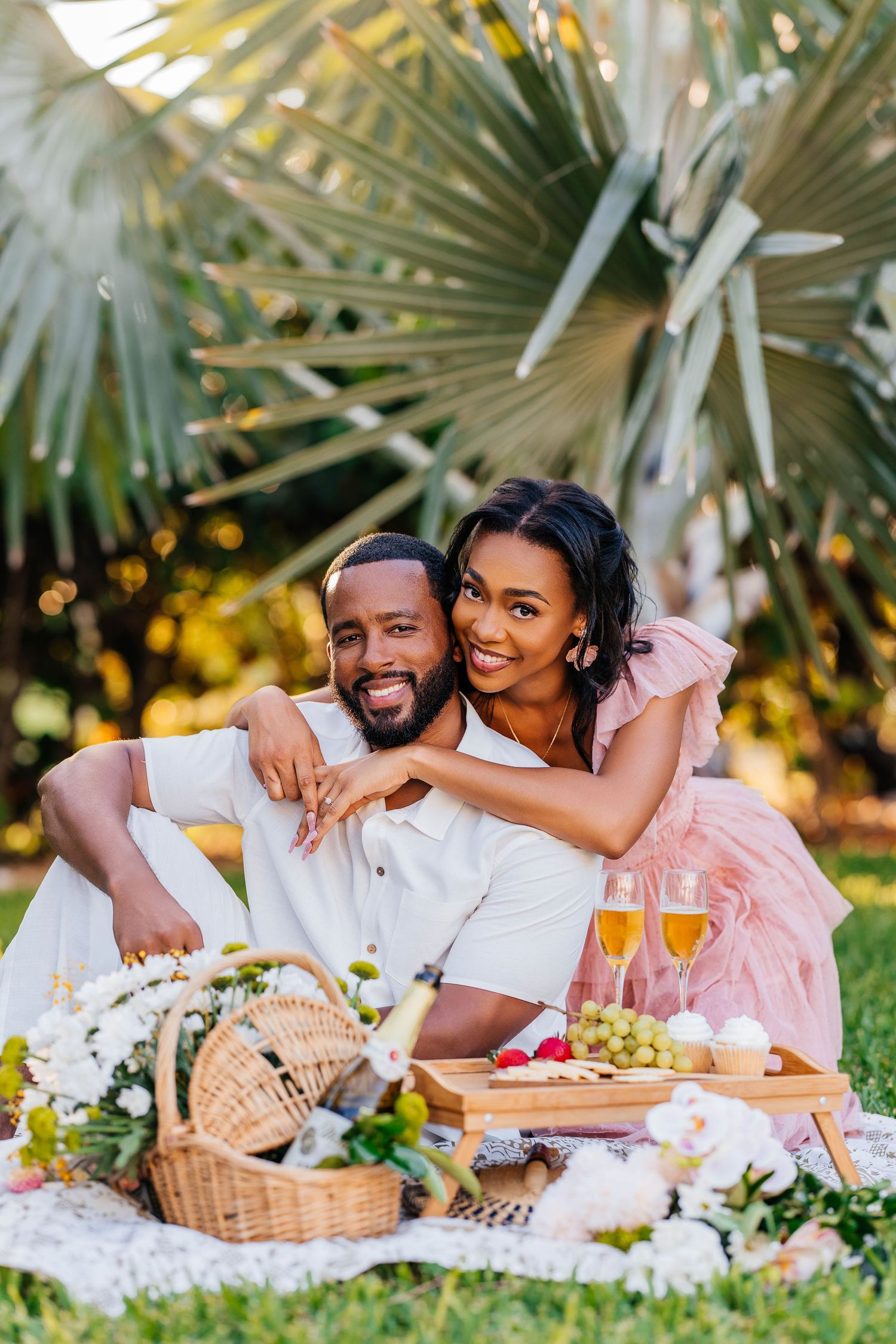 A couple enjoys a romantic outdoor picnic with wine and food baskets under tropical palm trees in a garden setting.