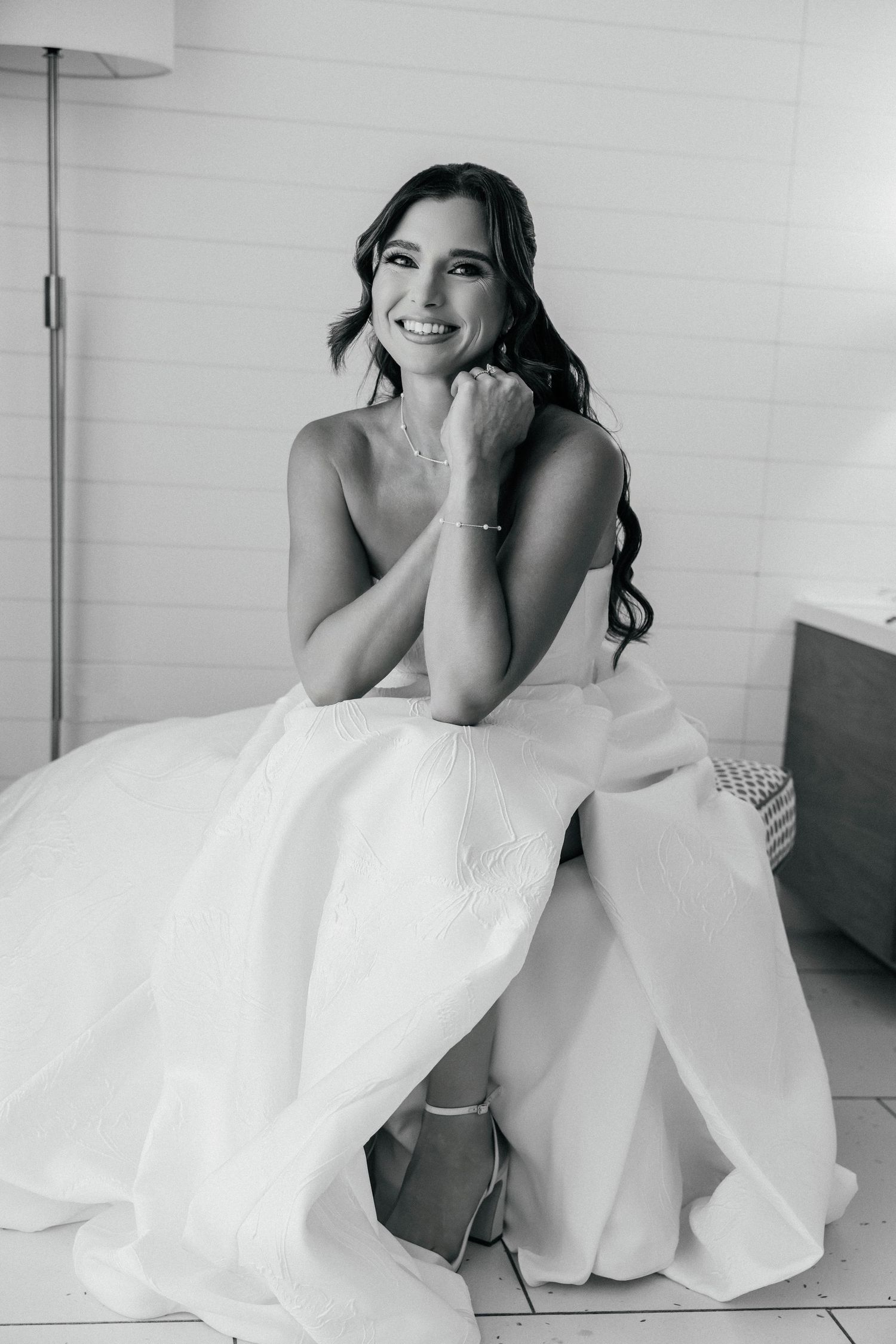 Black and white portrait of a smiling bride in a white gown, sitting elegantly with hands clasped near her chin.