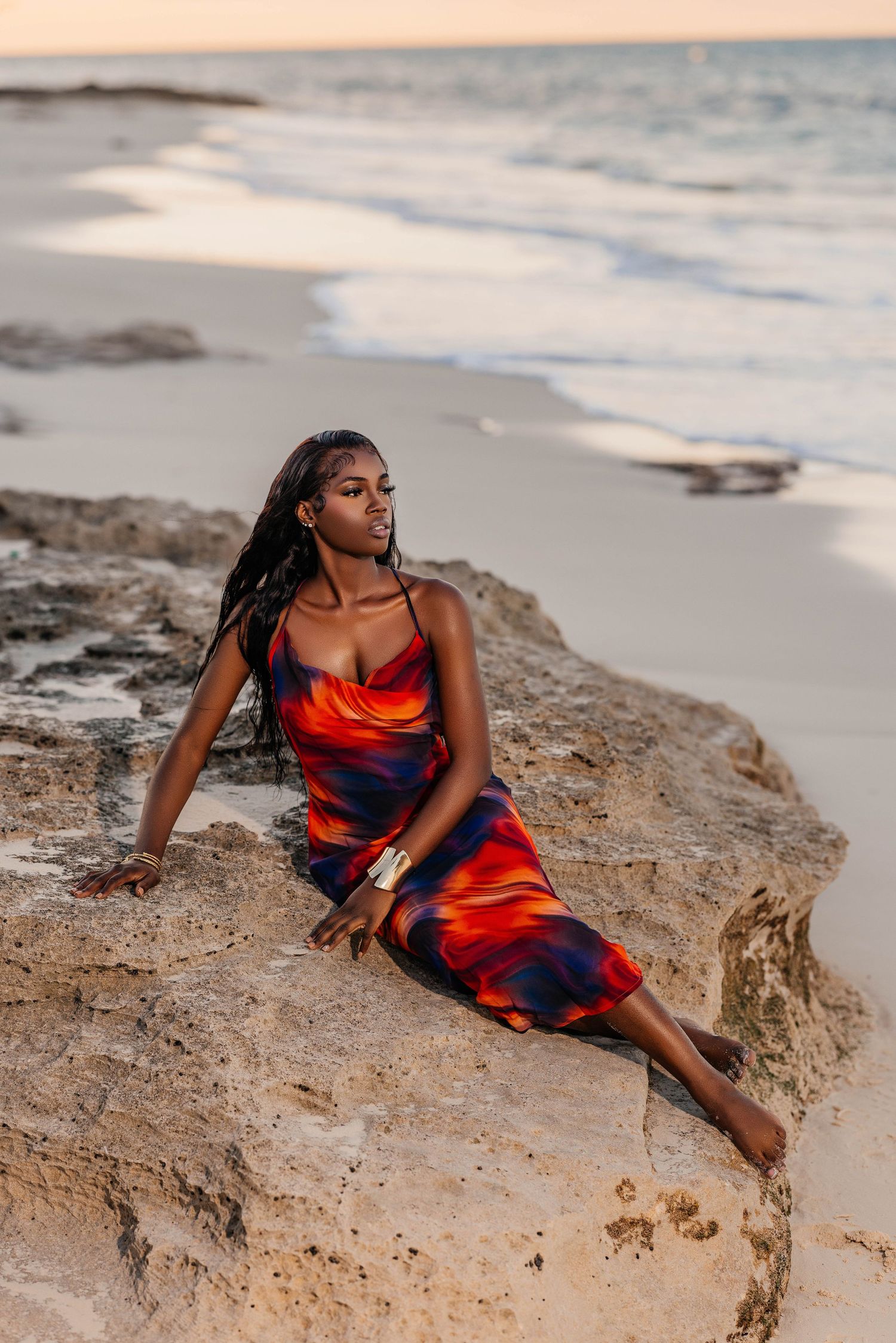 Woman in red and blue dress poses dramatically on beach rocks at sunset.