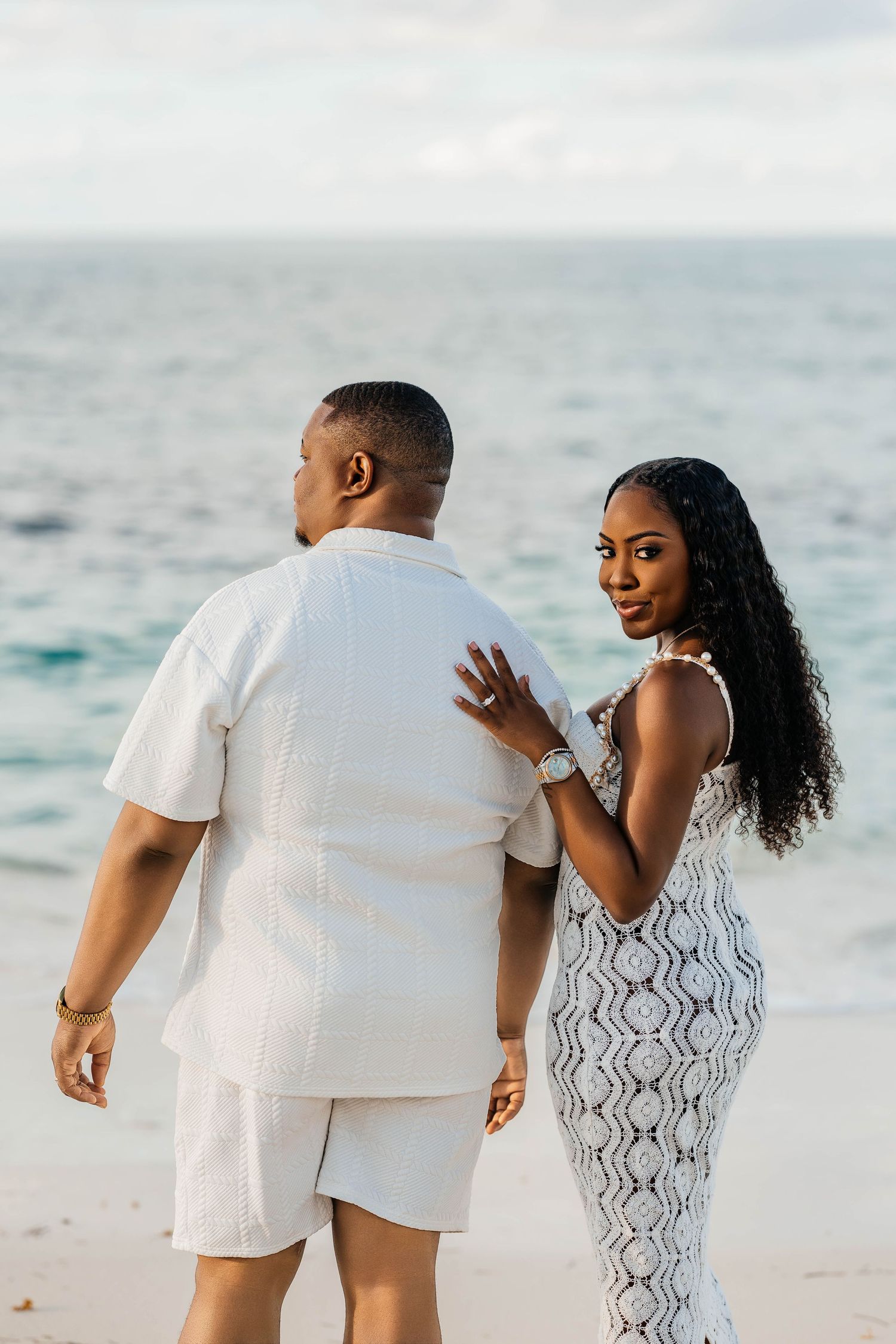 Couple enjoying a romantic moment on a beach wearing white outfits against an ocean backdrop.