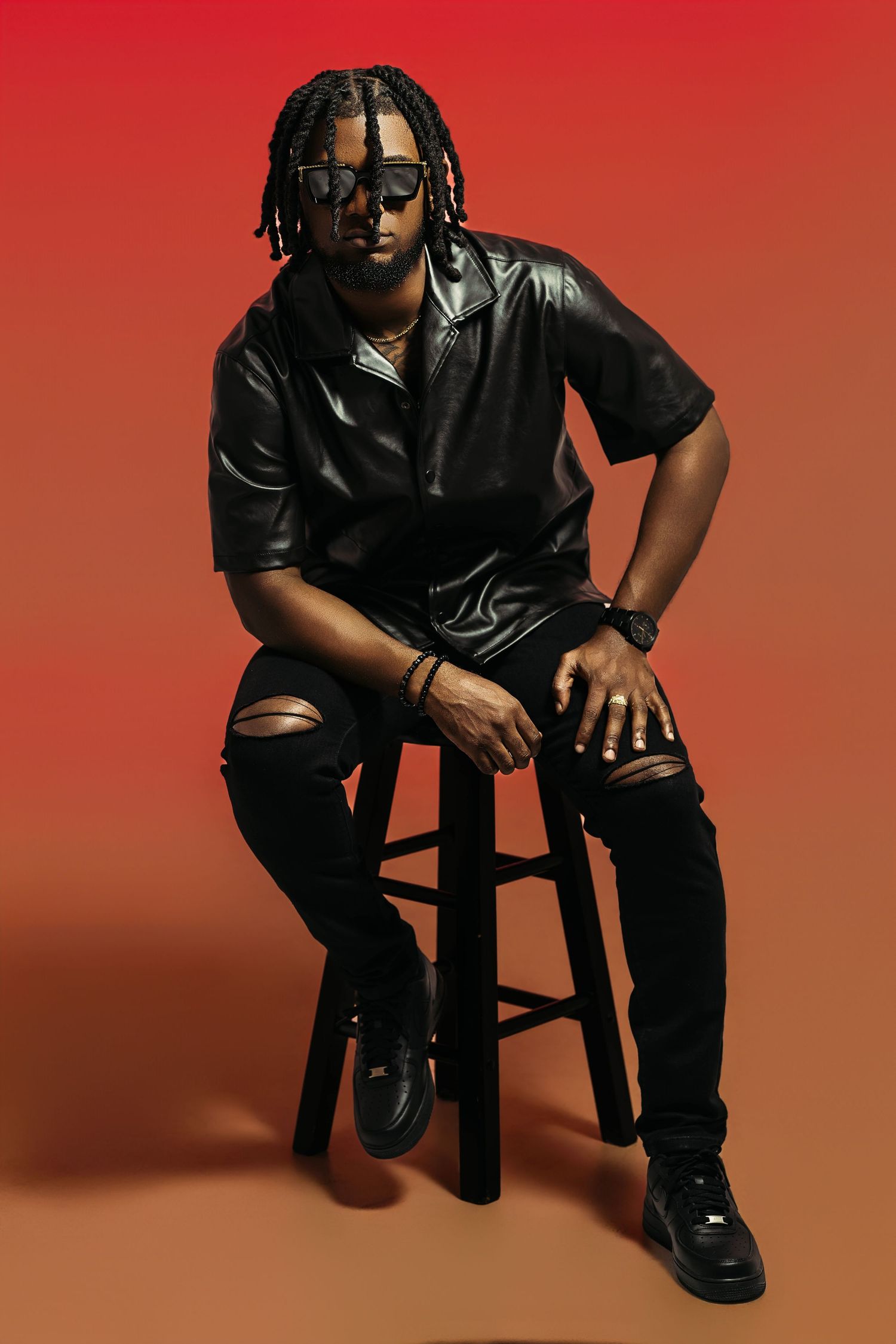 Person in all black attire poses on chair against red backdrop in artistic portrait series.