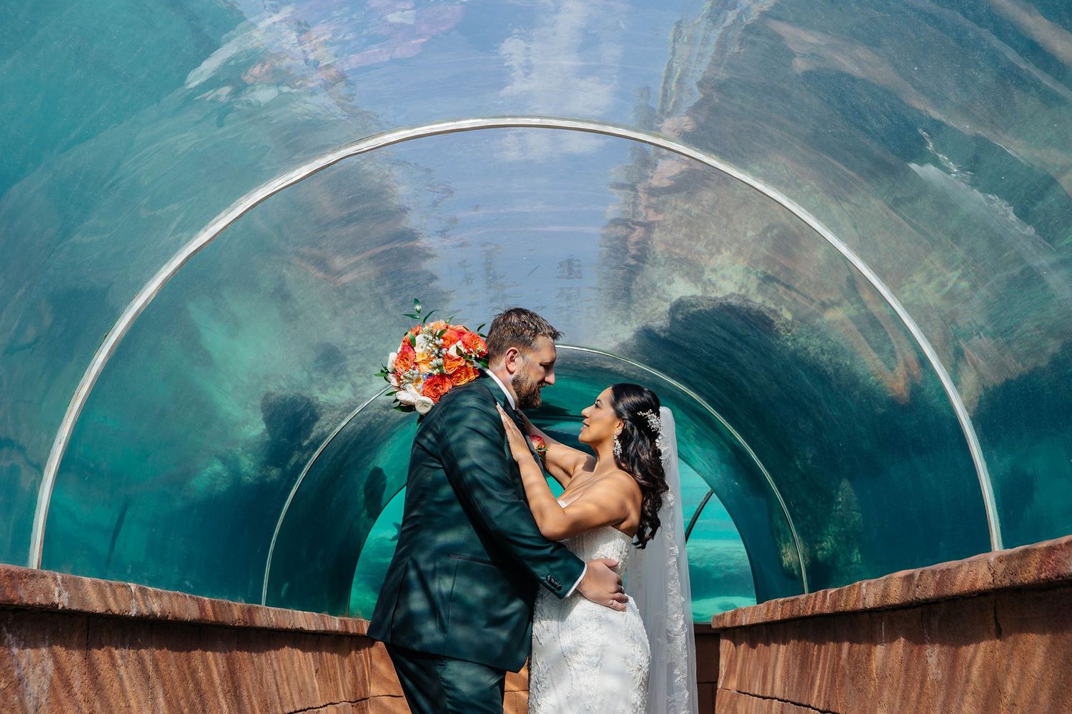 Romantic wedding photo in an aquarium tunnel with bright coral bouquet and teal water backdrop.
