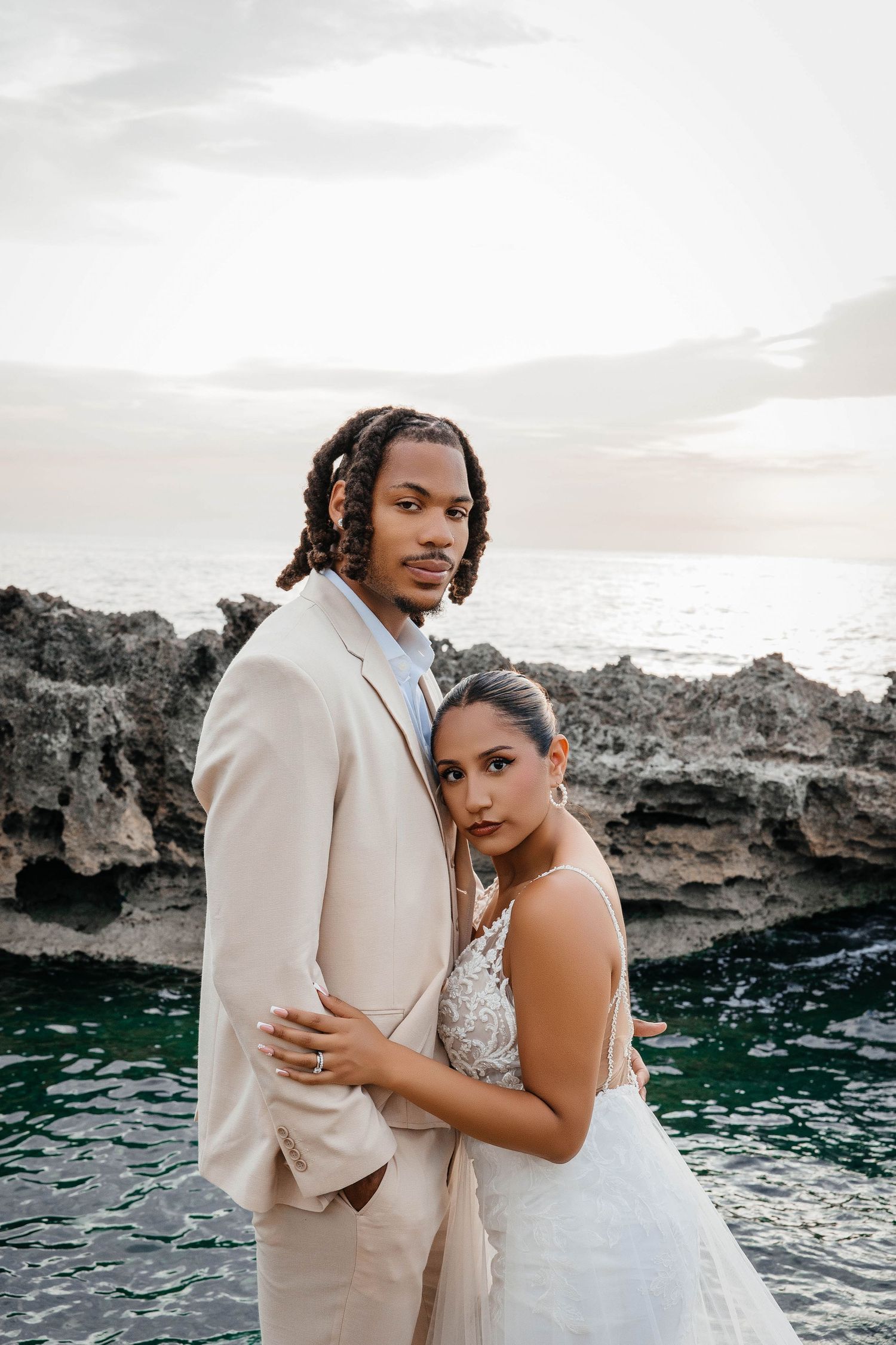 Romantic wedding portrait of couple embracing on rocky oceanside cliffs at sunset.