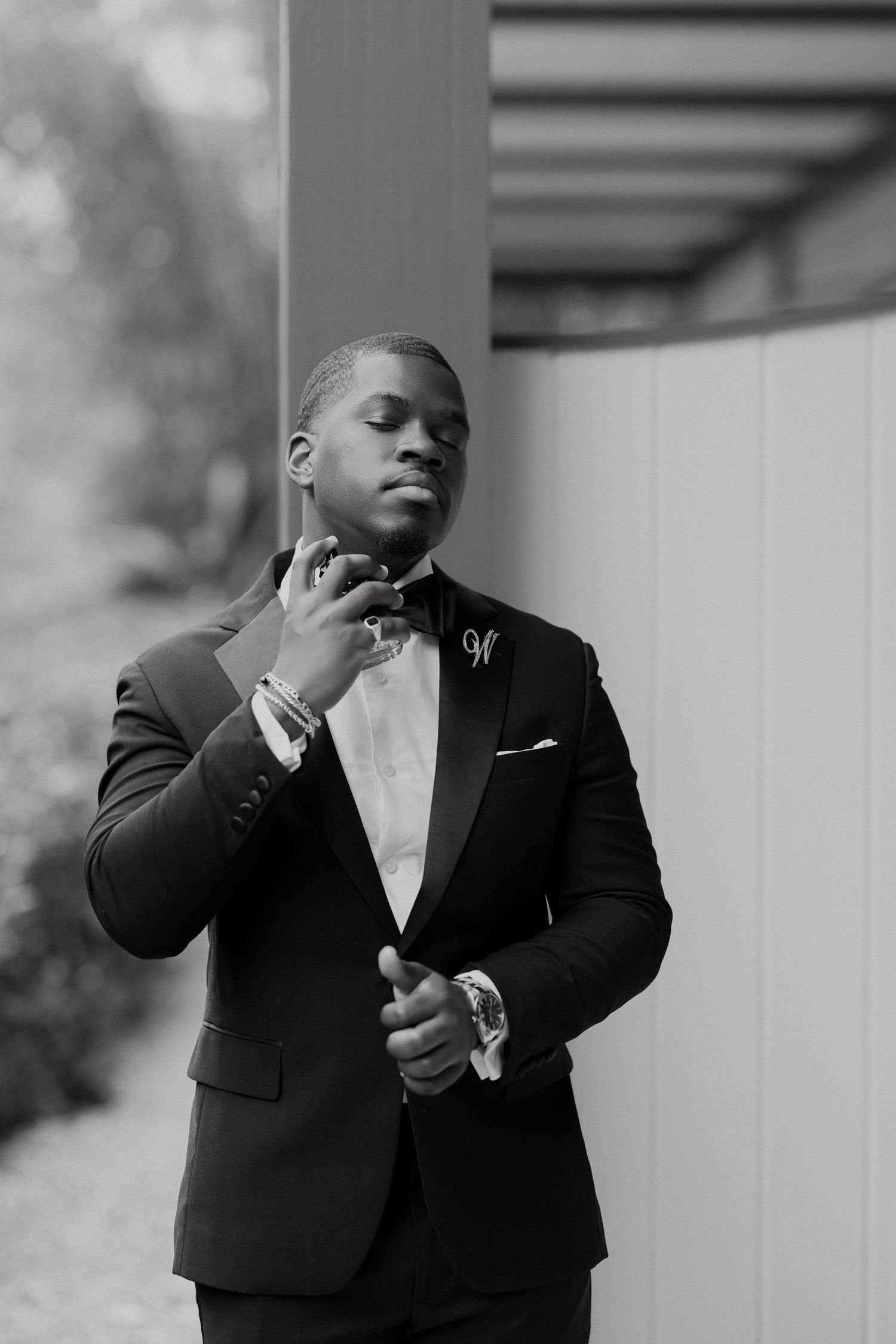 Black and white photo of a well-dressed groom in a tuxedo adjusting his bow tie before his wedding ceremony.