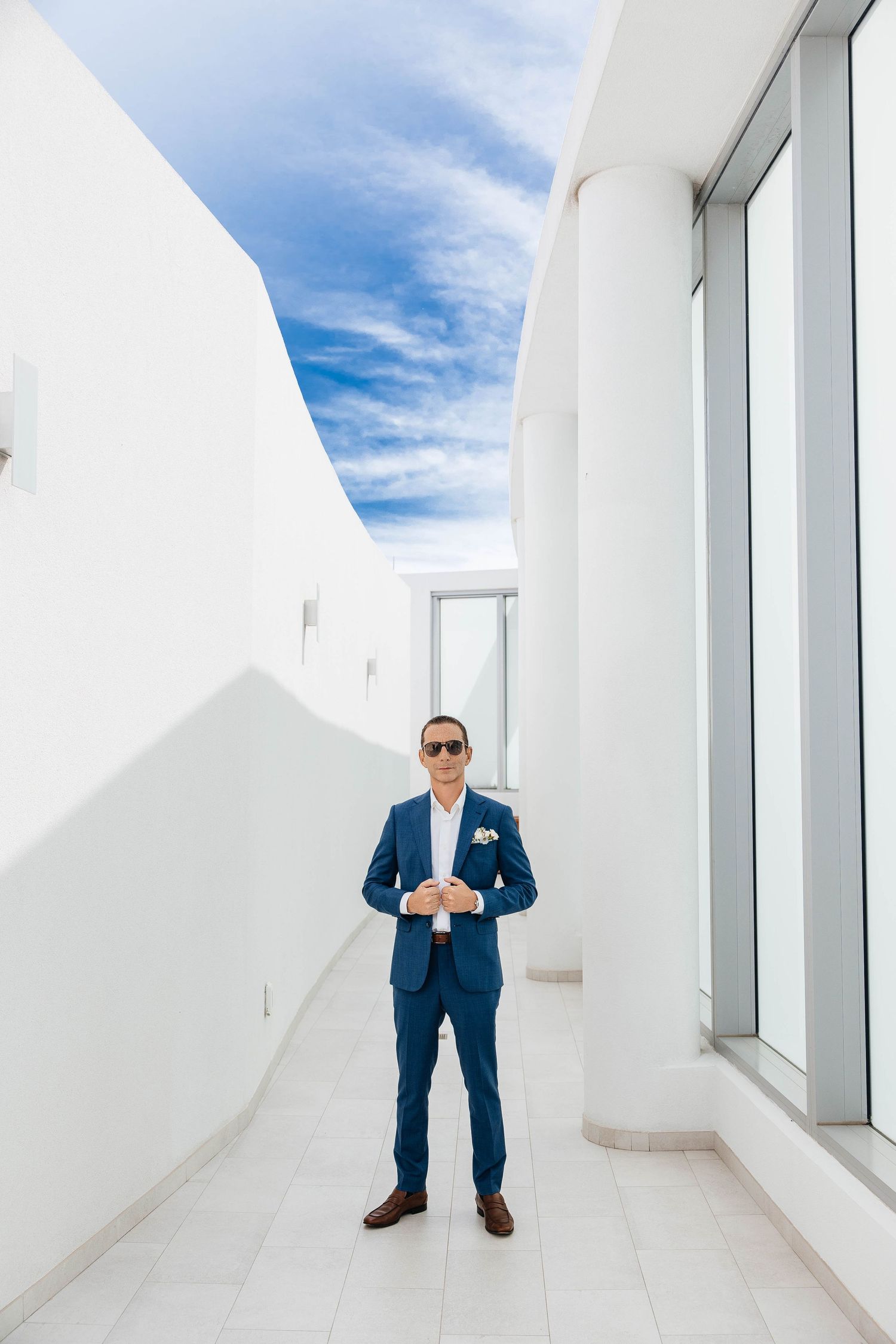 Groom in blue suit and sunglasses standing in white corridor with large windows and blue sky backdrop.