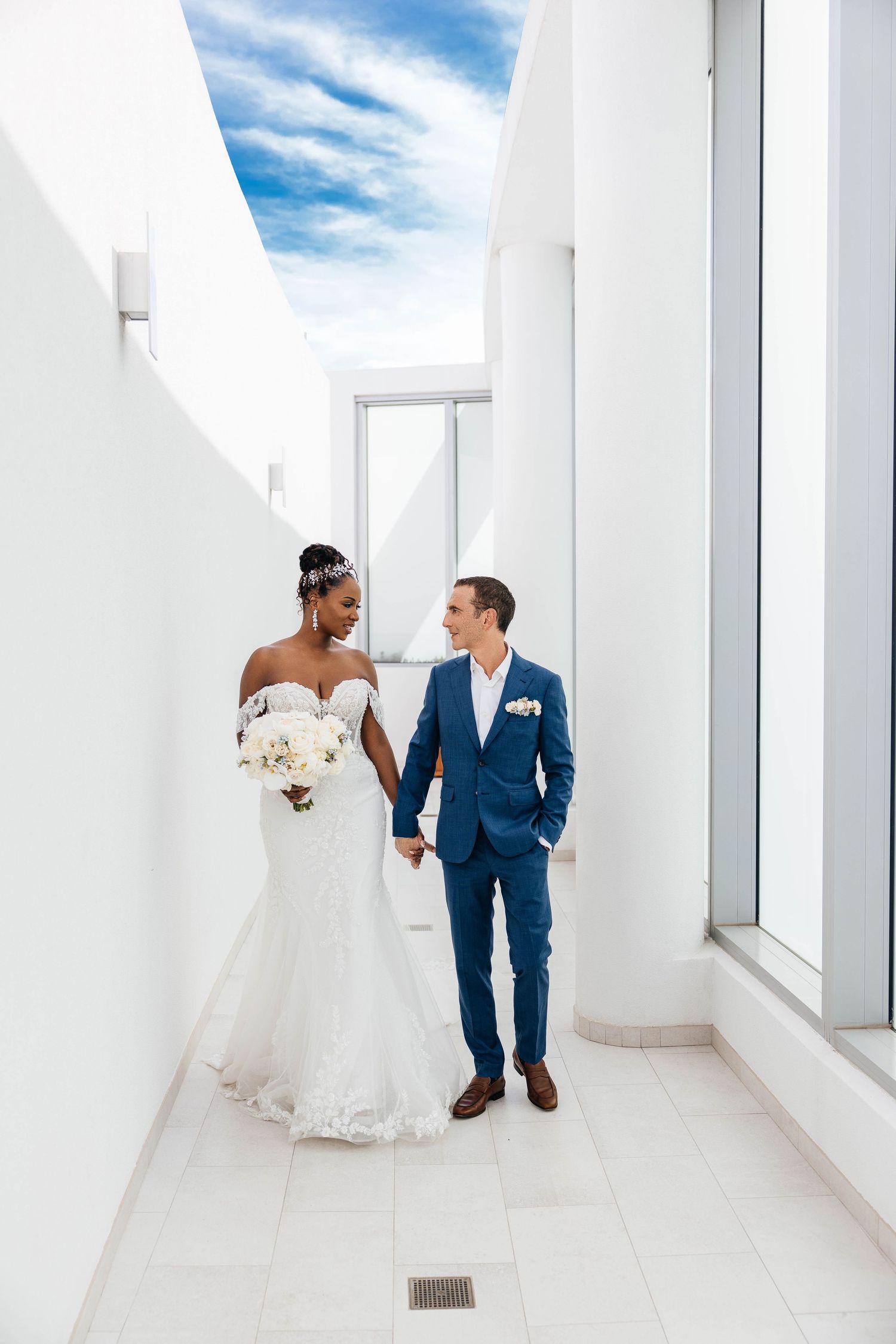 A bride and groom share a tender moment in a bright white hallway with floor-to-ceiling windows and blue sky views.
