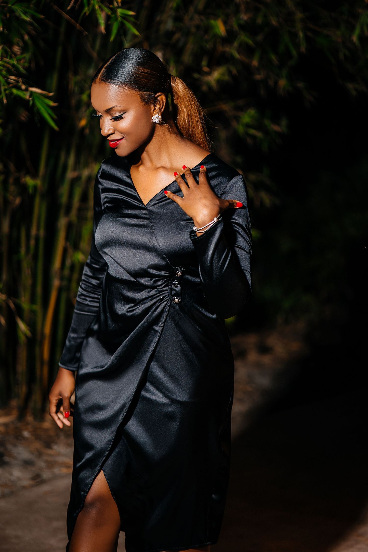 Woman in elegant black satin dress poses against dark bamboo garden background.
