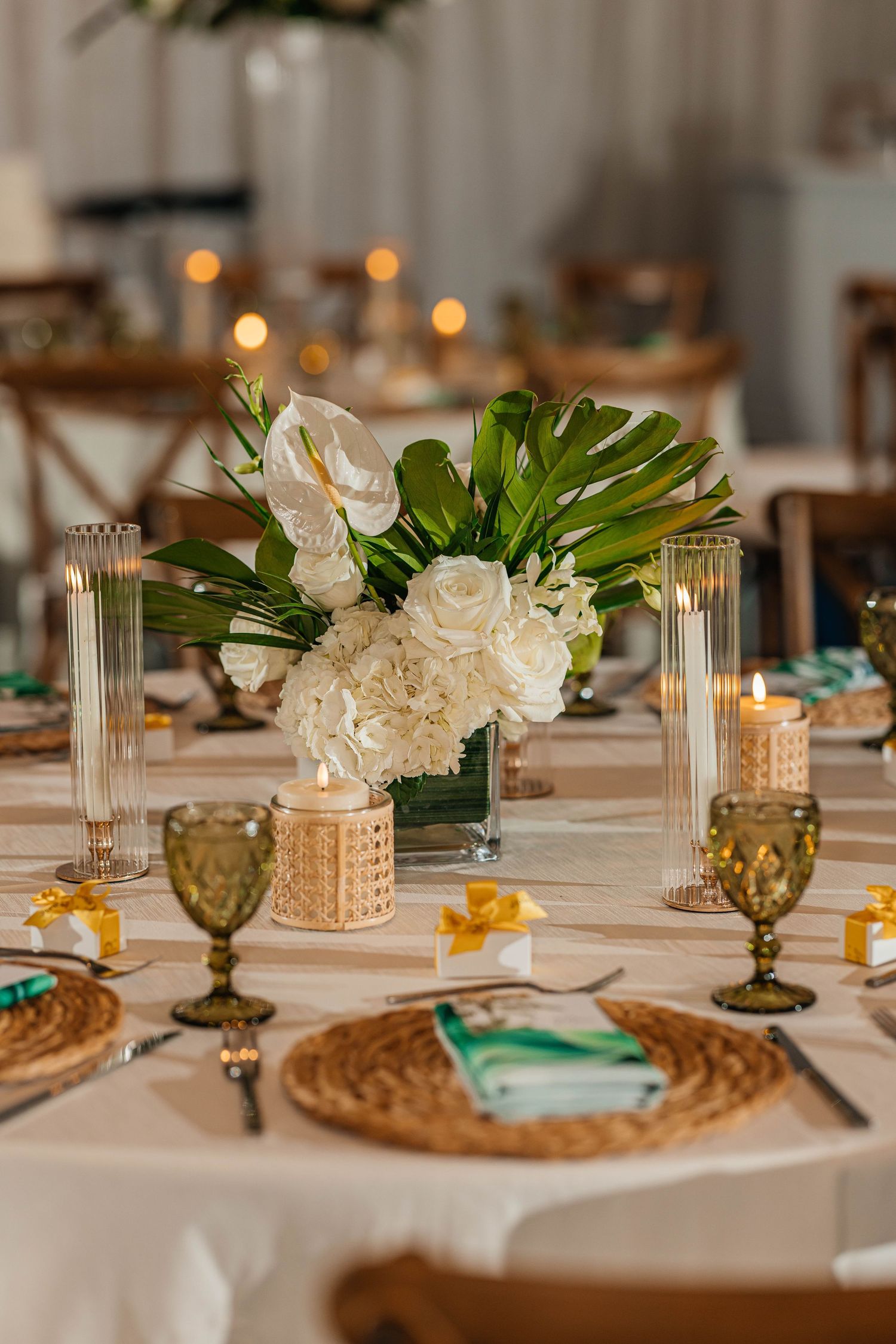 Elegant wedding table setting with white floral centerpiece, green tropical leaves, candles, and woven placemats.