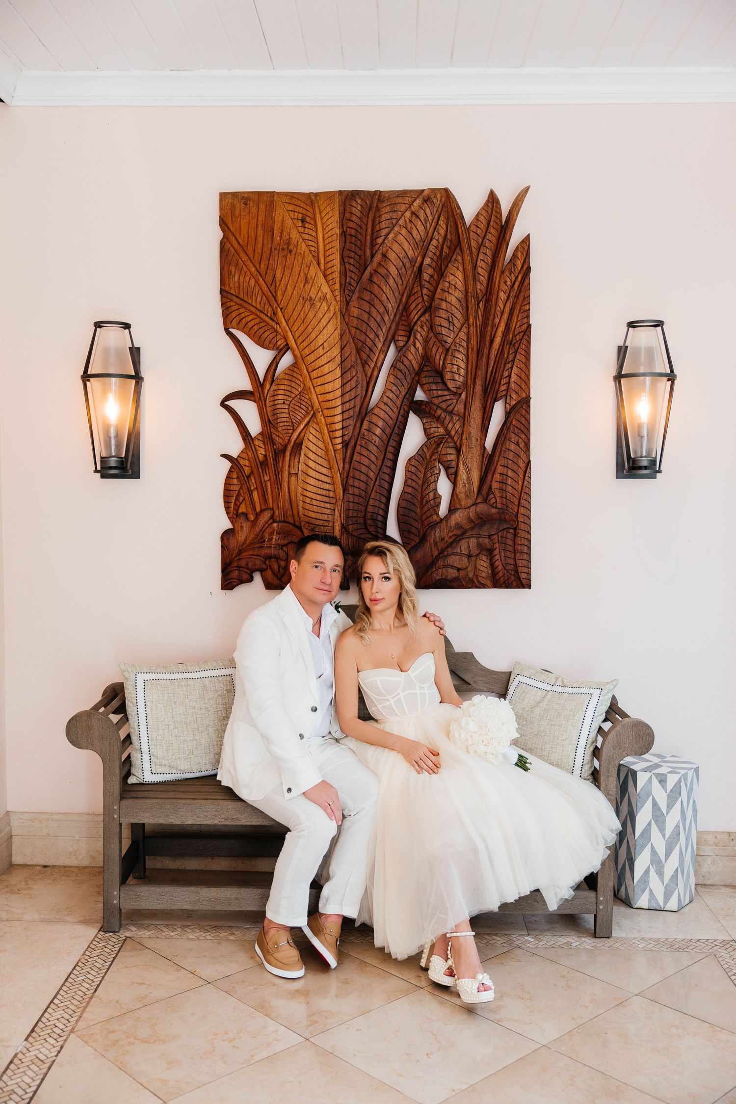Romantic tropical wedding setting with carved wooden palm leaf wall art and modern sconces above a cushioned bench.