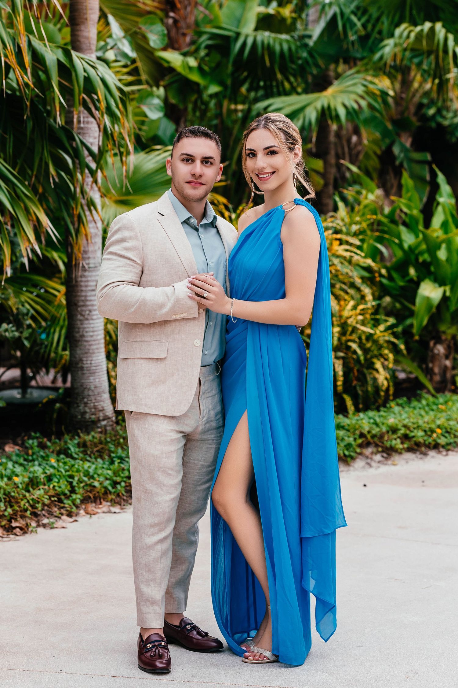 Elegant couple poses in tropical garden setting wearing beige suit and flowing blue dress.