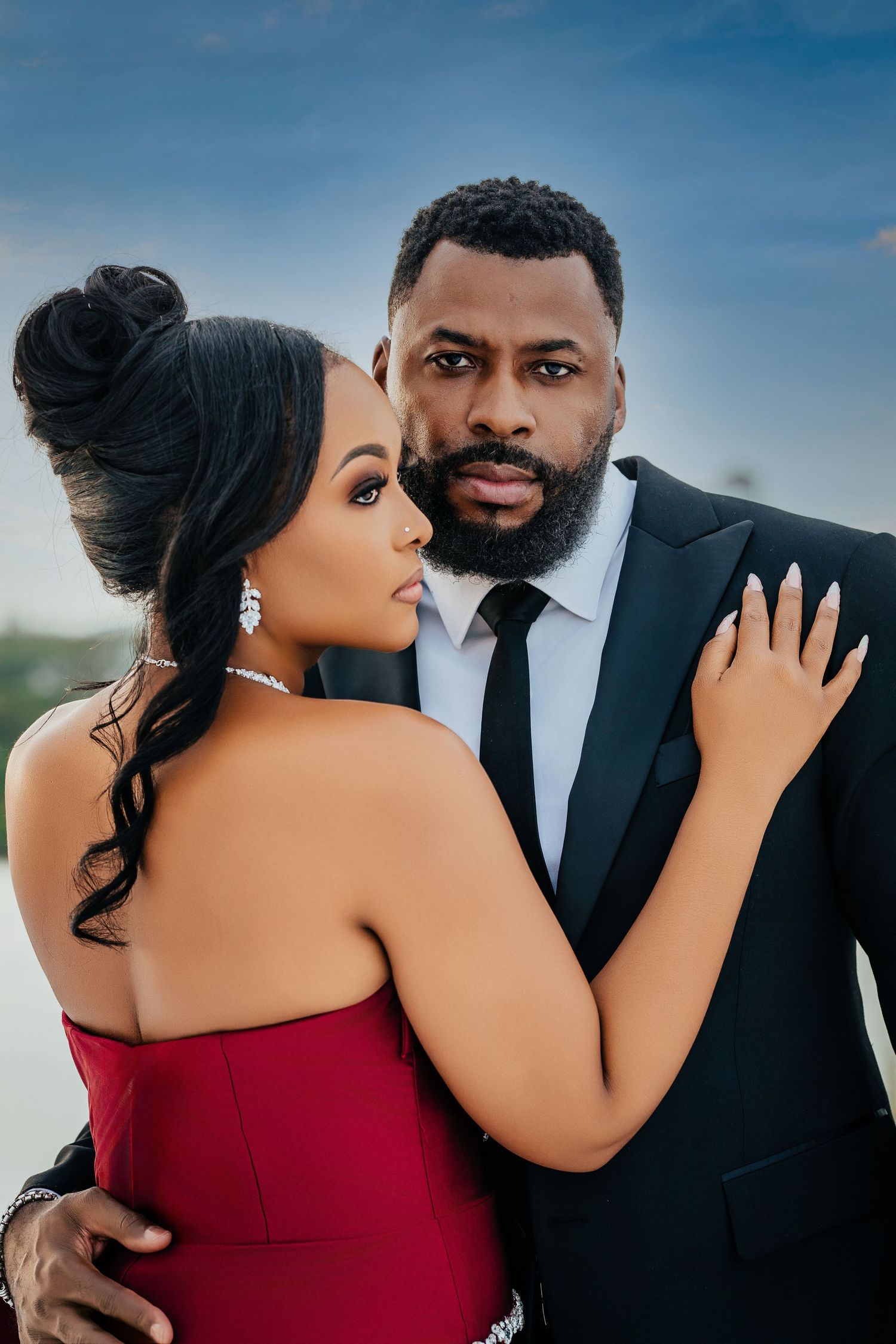 Romantic couple embraces in formal attire against blue sky backdrop during wedding photoshoot.