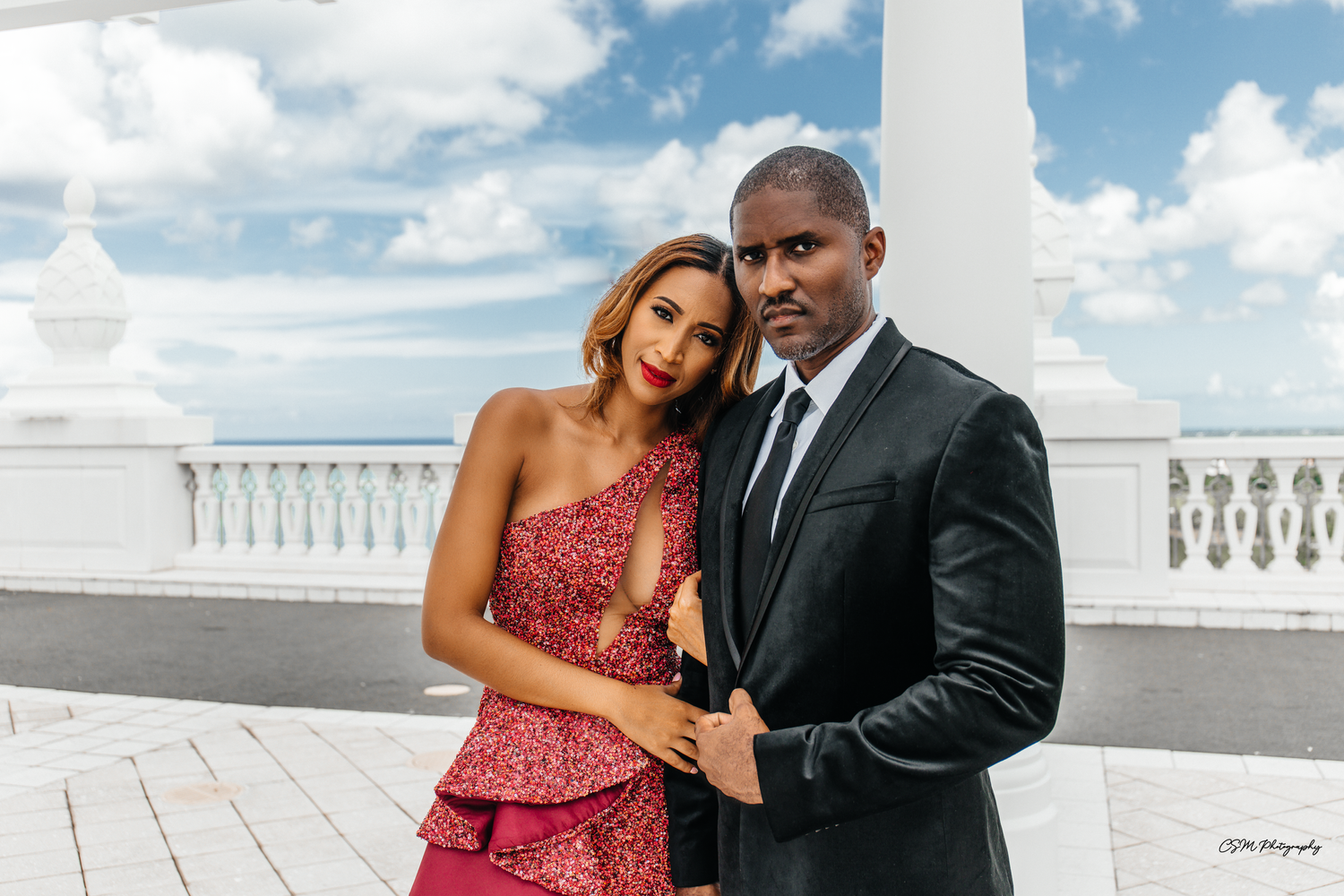 Elegant couple poses on white balcony under bright blue sky.