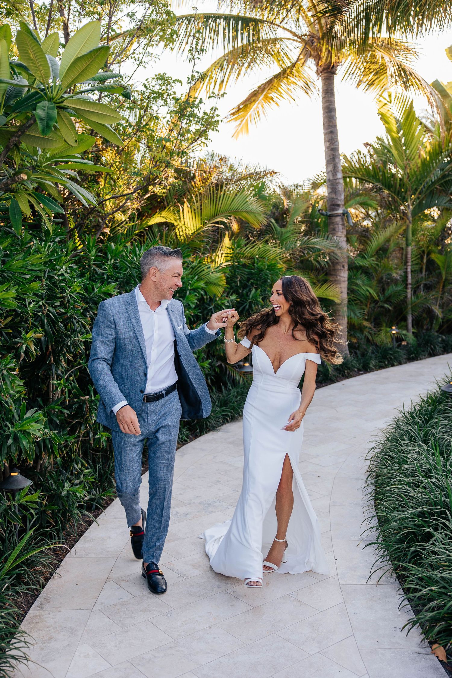 Wedding couple holding hands while walking down scenic palm-lined pathway.