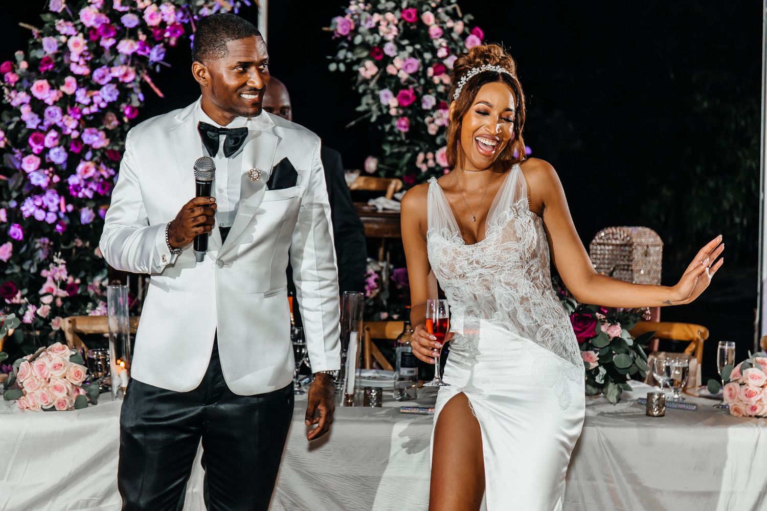 Bride and groom laughing together at wedding reception, bride in white gown holding wine glass, groom in white tuxedo jacket.