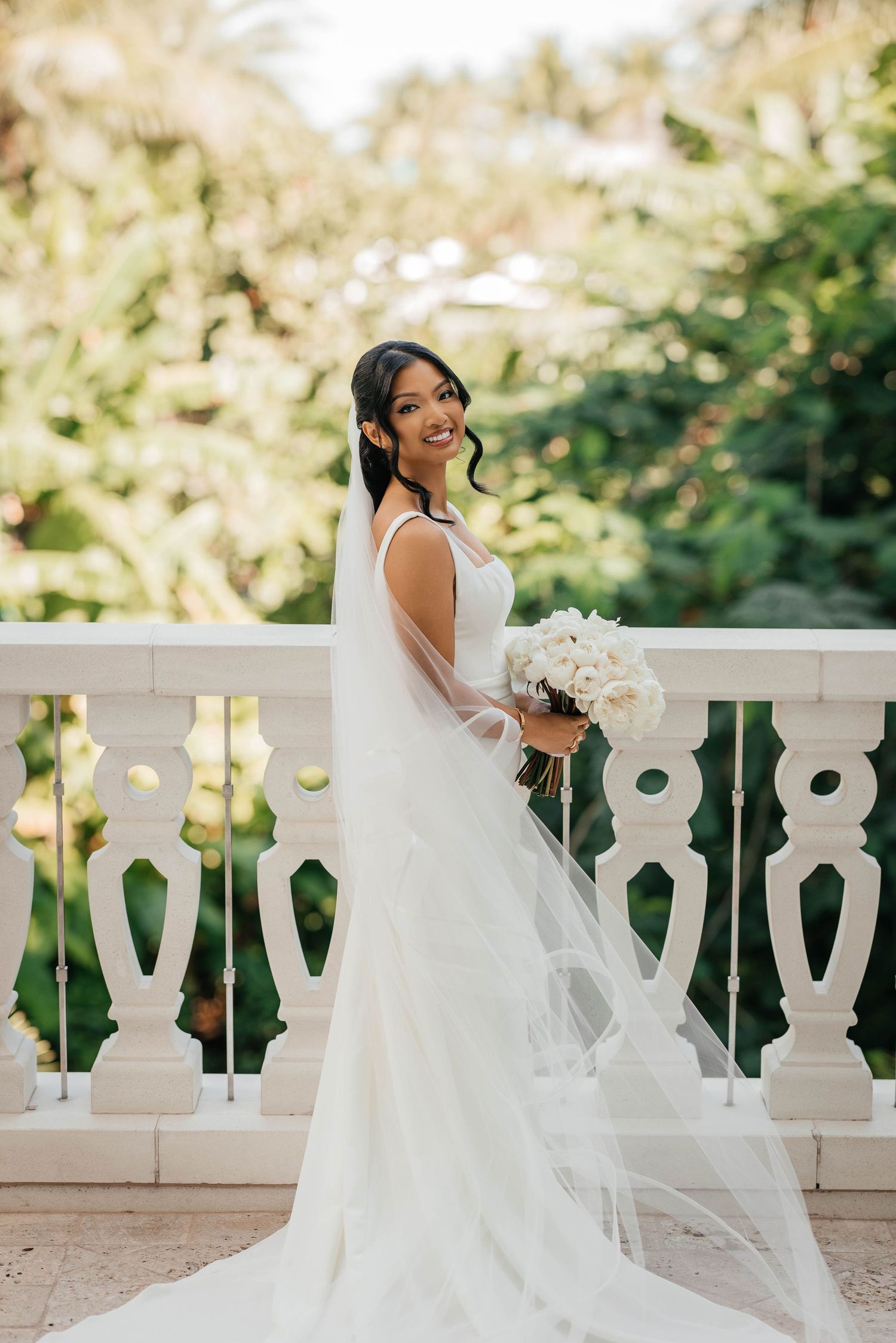A bride in a flowing white wedding gown sits gracefully on an ornate stone balustrade in a sunlit garden setting.