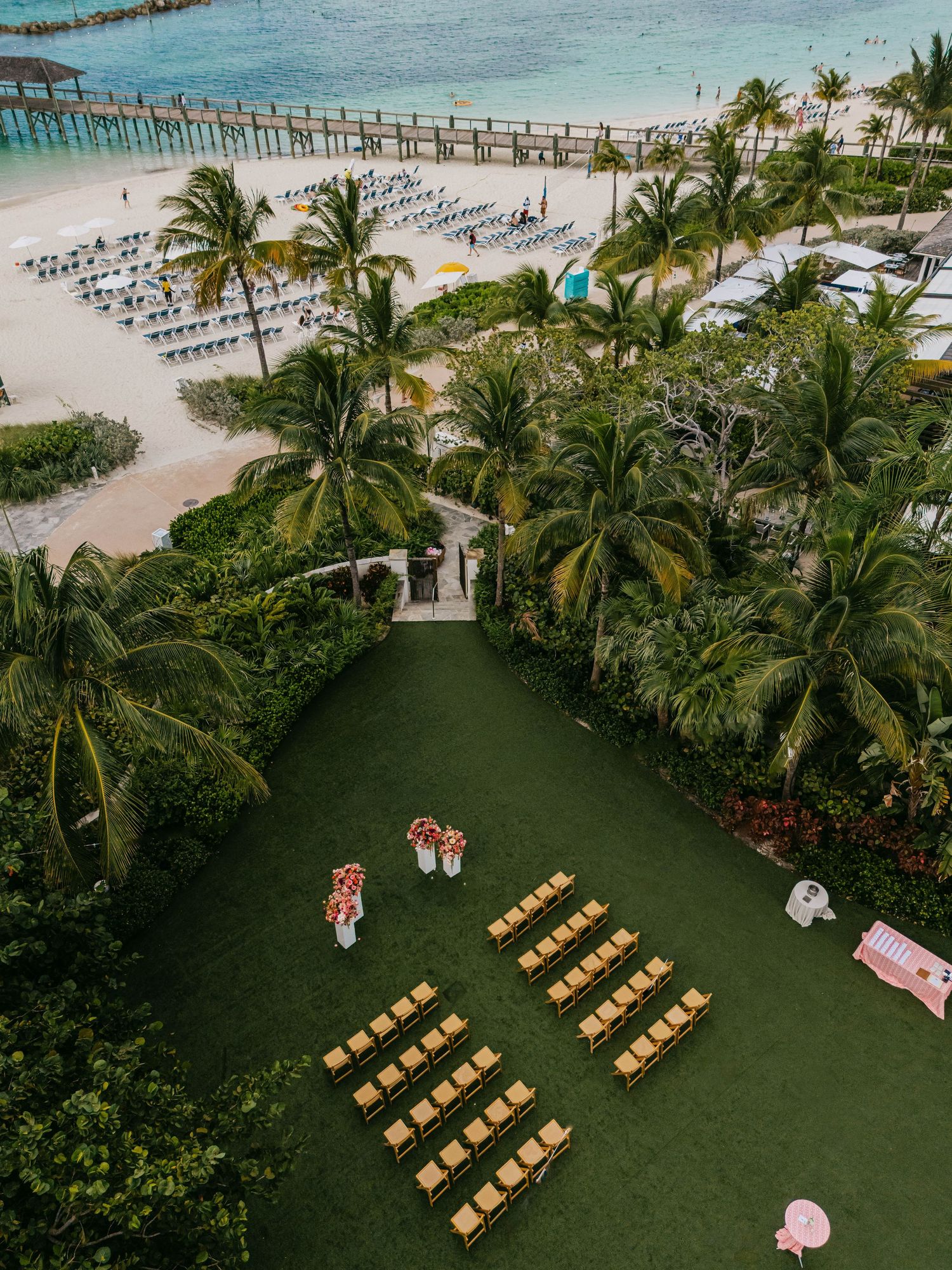 Aerial view of a tropical beach wedding ceremony setup with rows of chairs on green lawn, palm trees, and ocean backdrop.