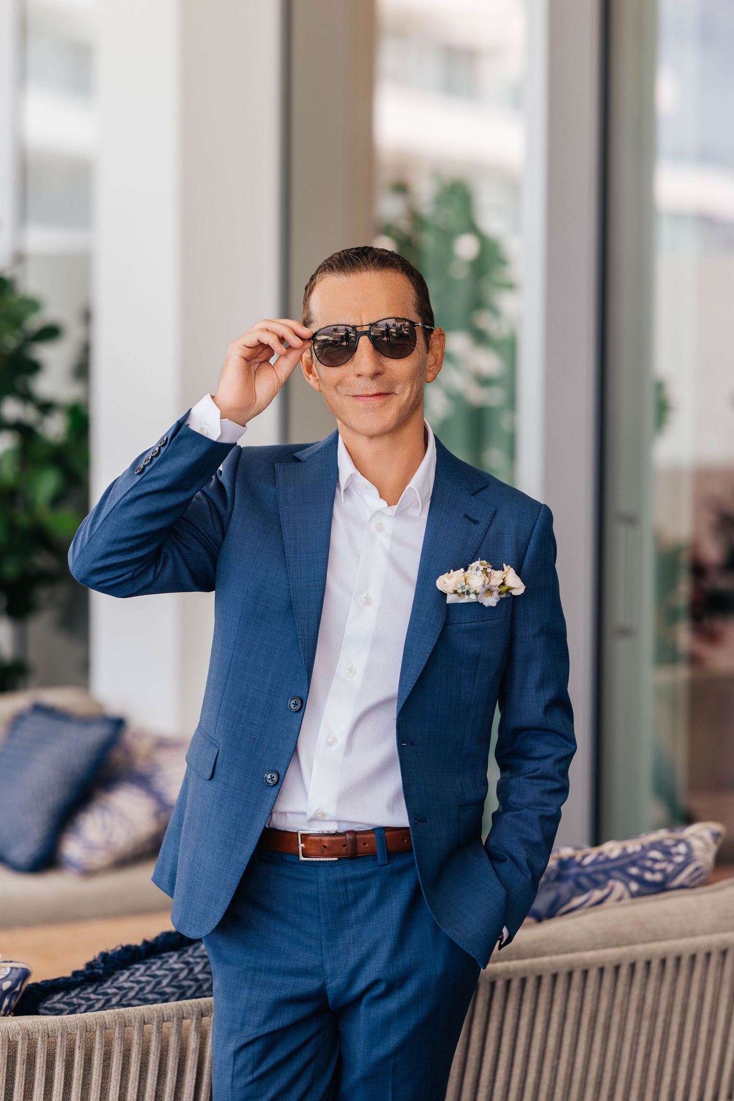 Stylish man in blue suit adjusting sunglasses, wearing white shirt with floral pocket square, standing indoors.