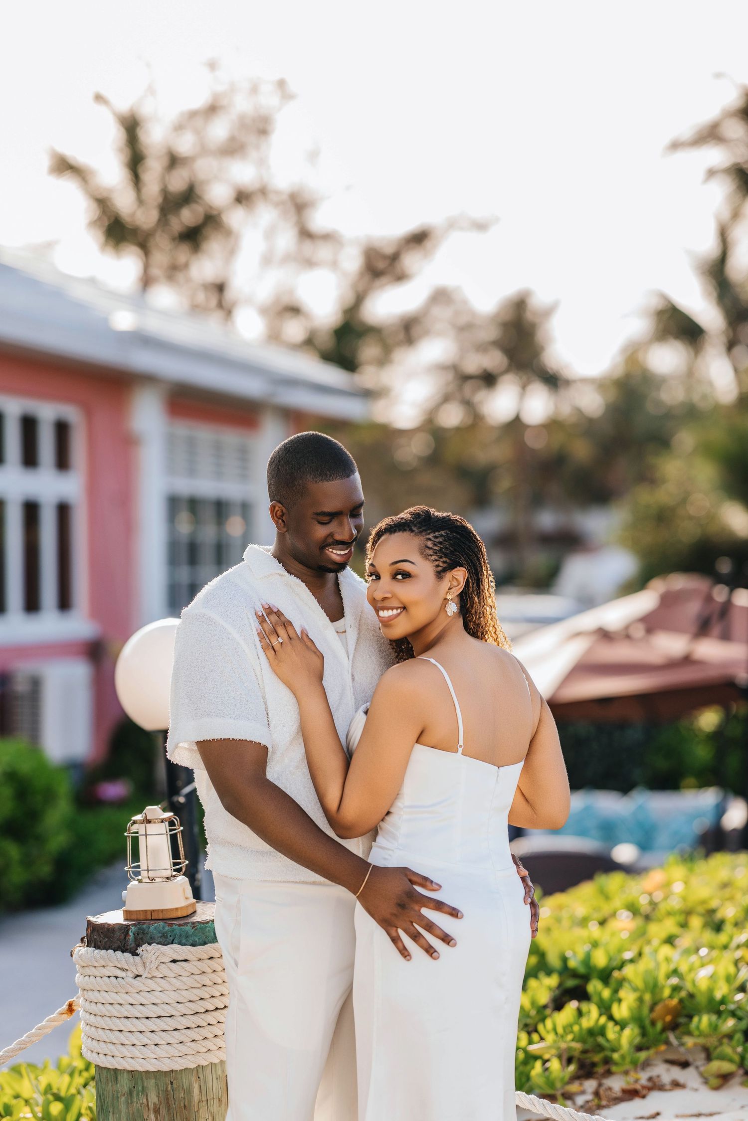 A couple in white outfits embrace during a romantic outdoor photoshoot near tropical palm trees and pink buildings.