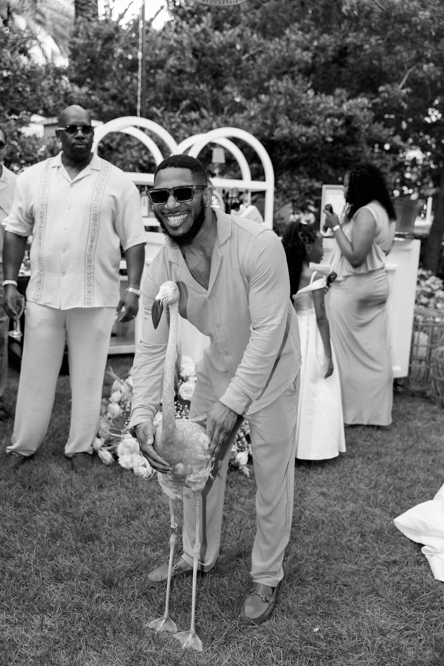 Man in sunglasses smiling while posing with a flamingo at an outdoor wedding celebration in black and white.