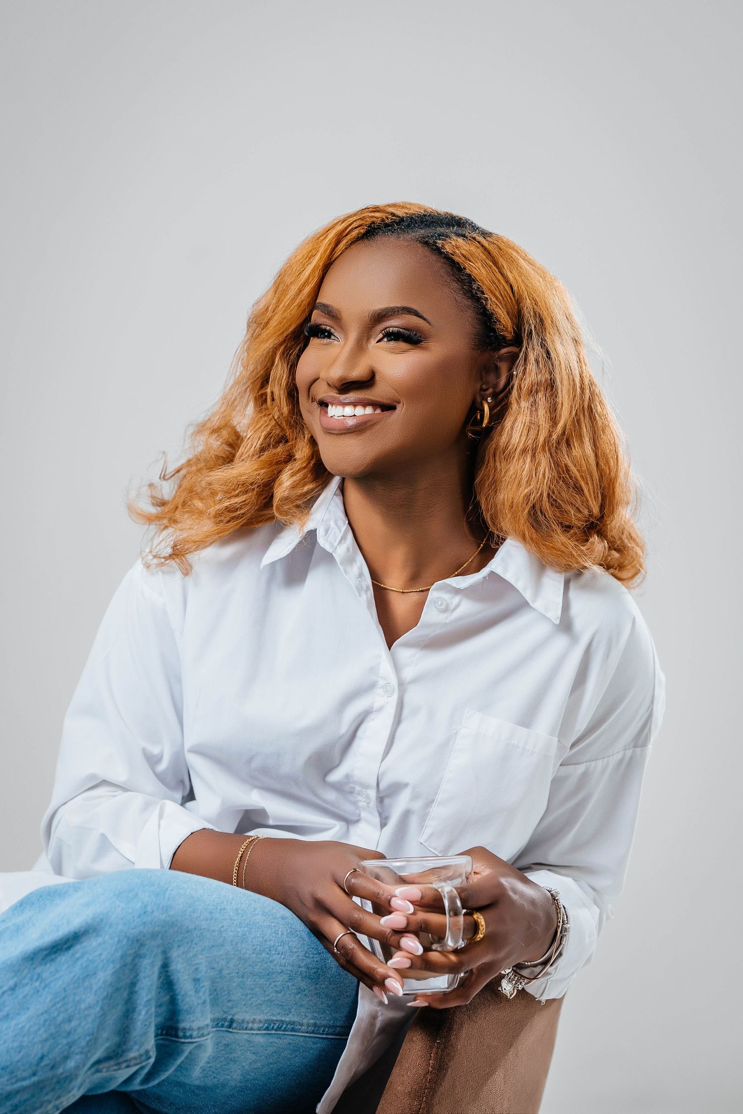 Person with wavy blonde hair wearing a white collared shirt smiling warmly in a studio setting.