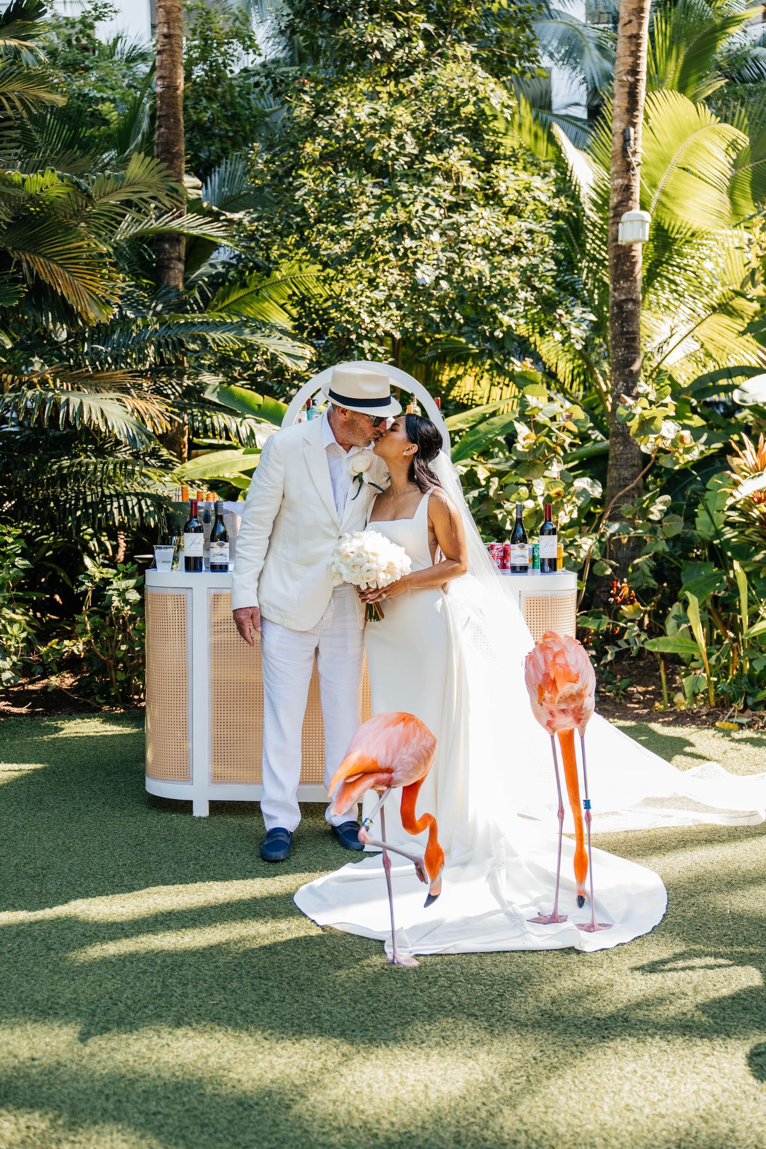 Bride and groom kissing at tropical wedding with pink flamingos, lush greenery, and a bar backdrop.