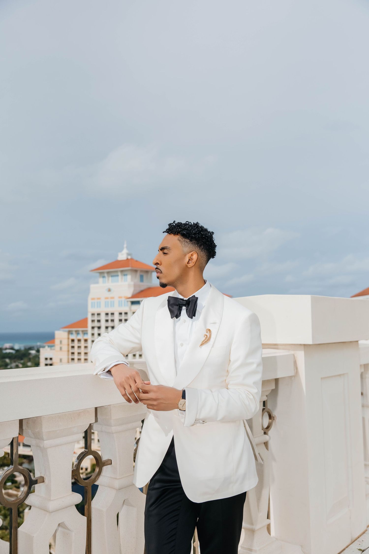 A well-dressed man in a white tuxedo jacket and black bow tie stands on a rooftop balcony with a city skyline behind him.