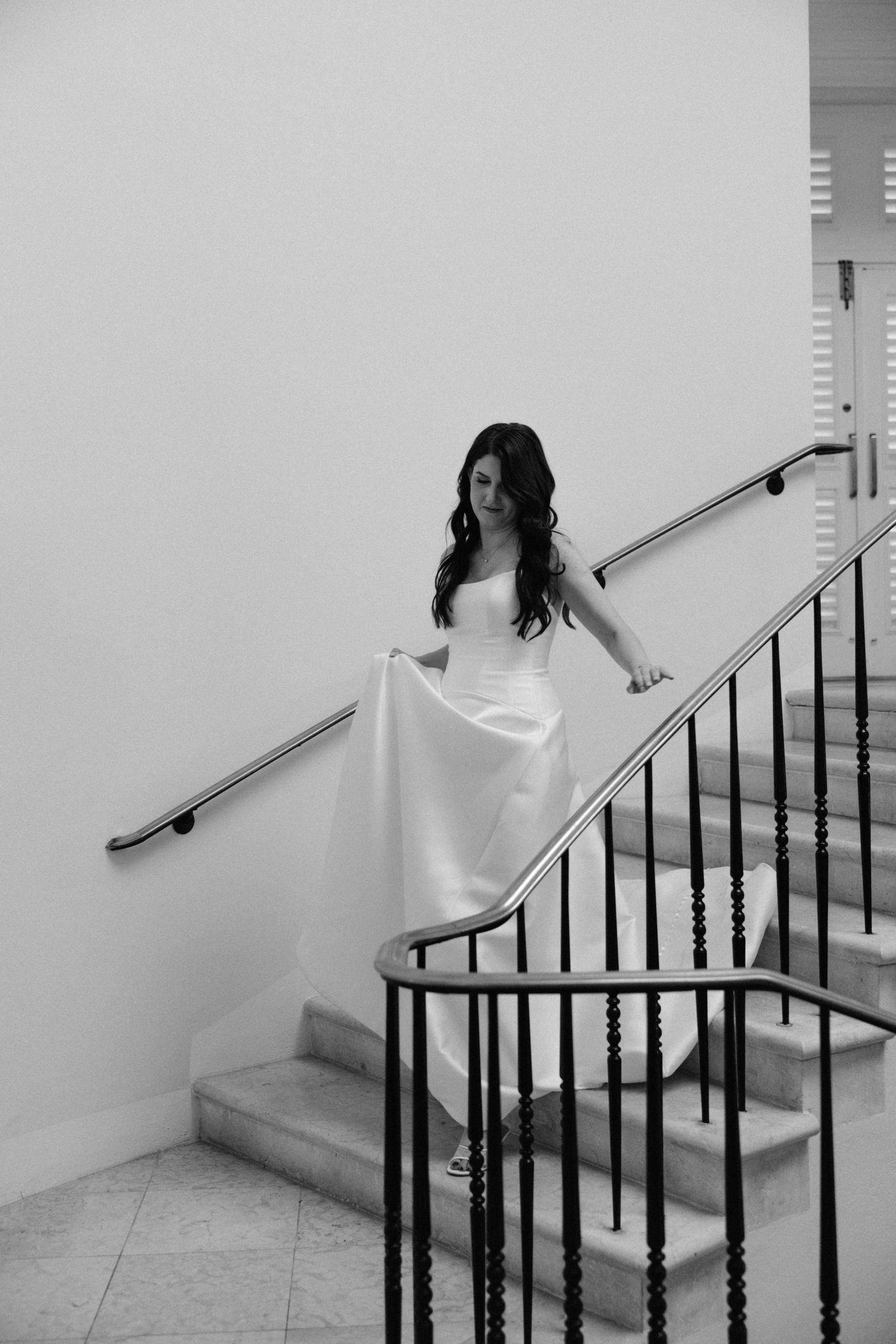 Bride in a white strapless gown descending an elegant staircase with black iron railings in a black and white photo.