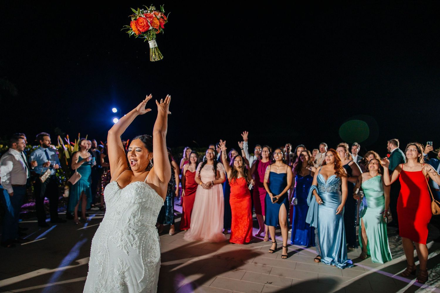 Bride tossing wedding bouquet to excited guests in colorful dresses at an outdoor nighttime wedding reception.