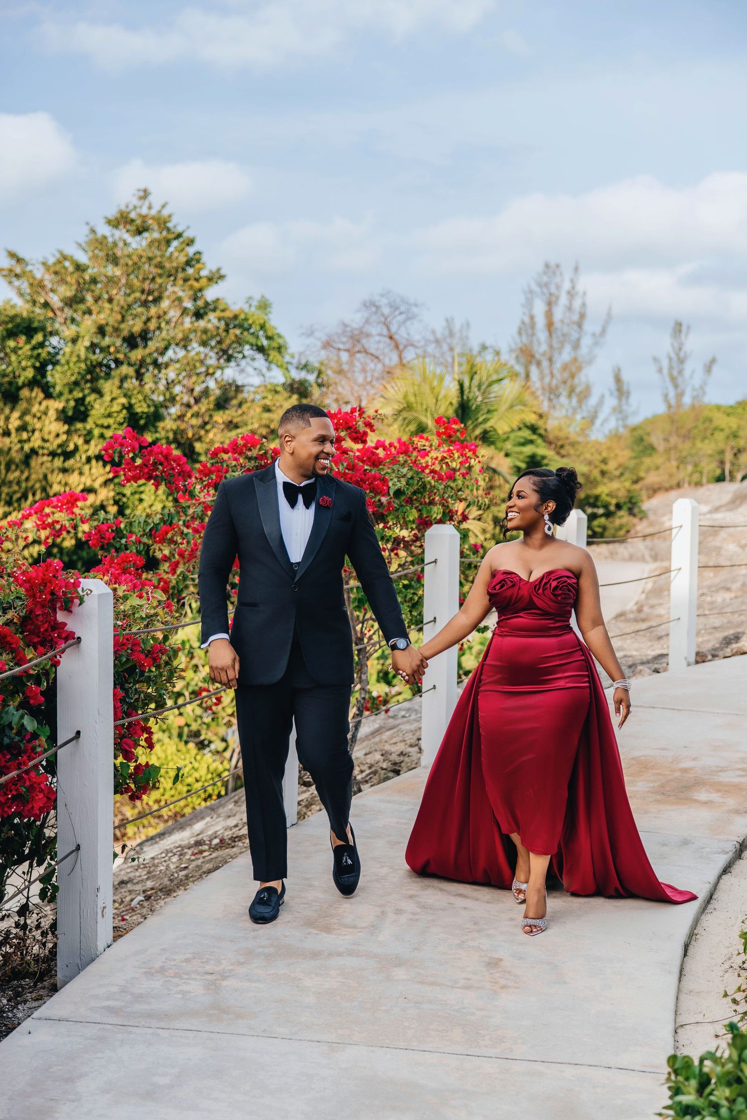 Couple in formal attire walking along garden path with blooming red flowers.