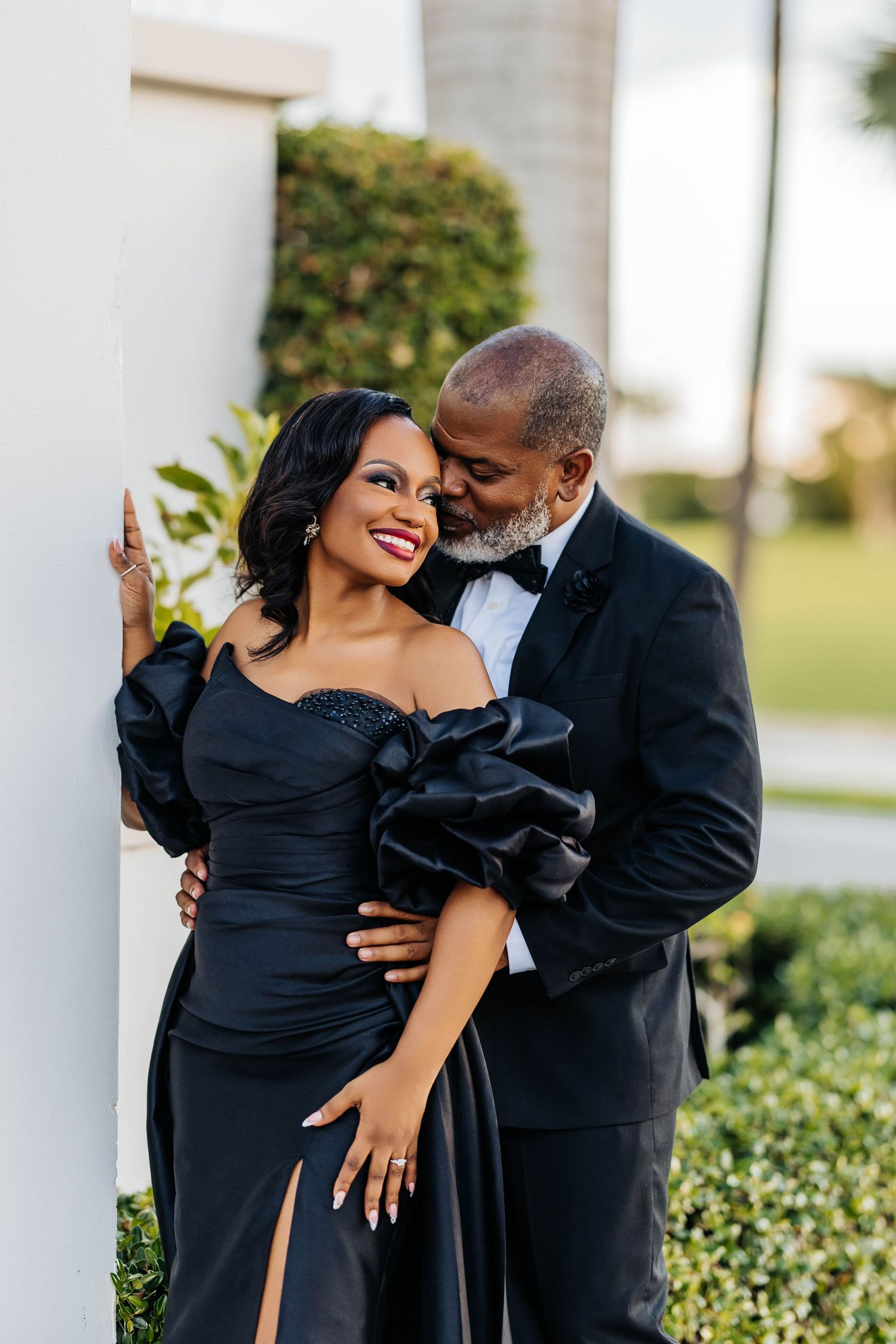 A couple in formal black attire embraces romantically in a series of outdoor photos near white columns and greenery.