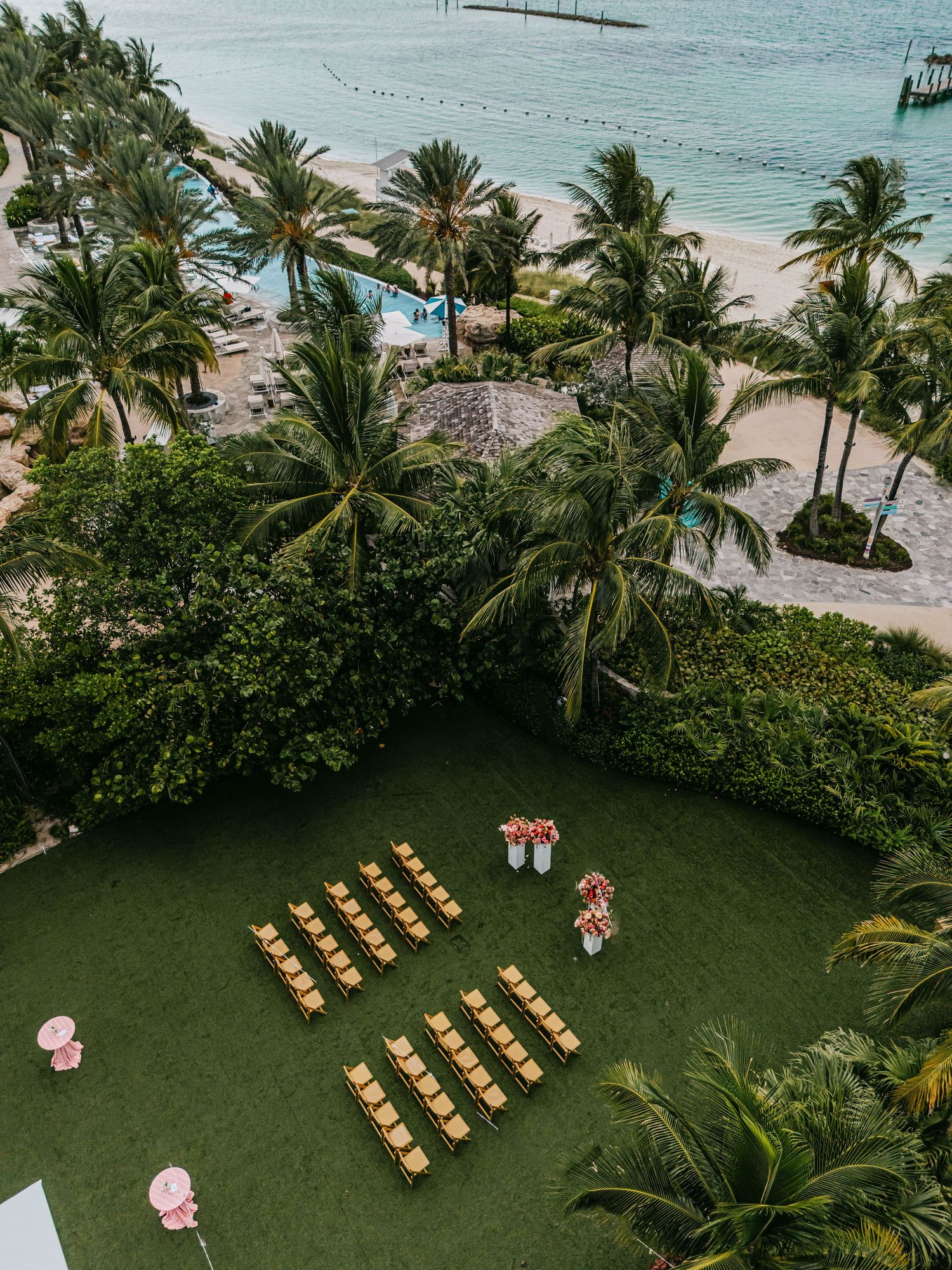 Aerial view of an outdoor beach wedding setup with gold chairs on green lawn, palm trees, and turquoise ocean backdrop.