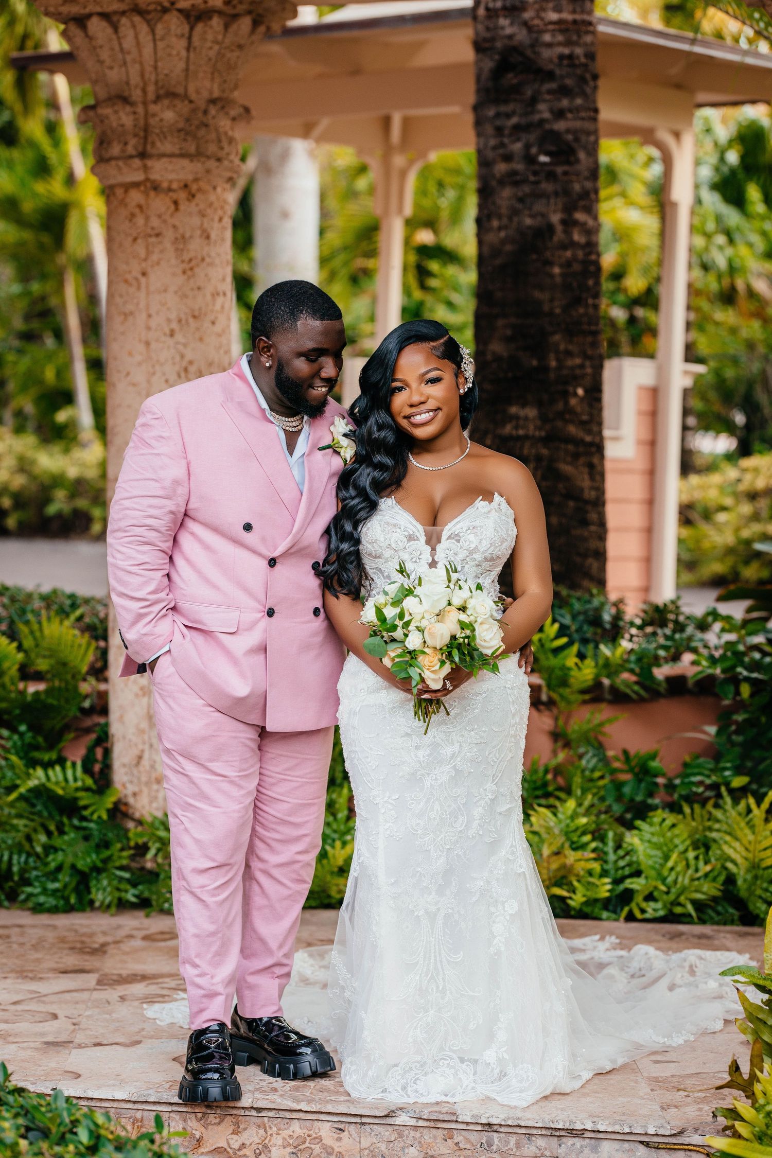 Elegant wedding portrait with pink double-breasted suit and white lace gown in tropical setting.