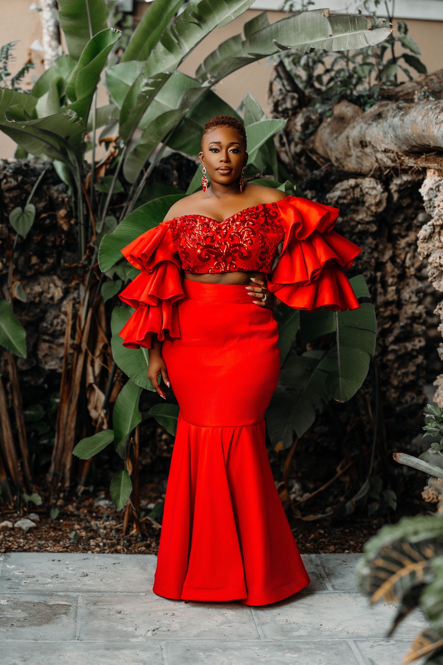 Elegant red two-piece formal dress with ruffled sleeves against tropical garden backdrop.