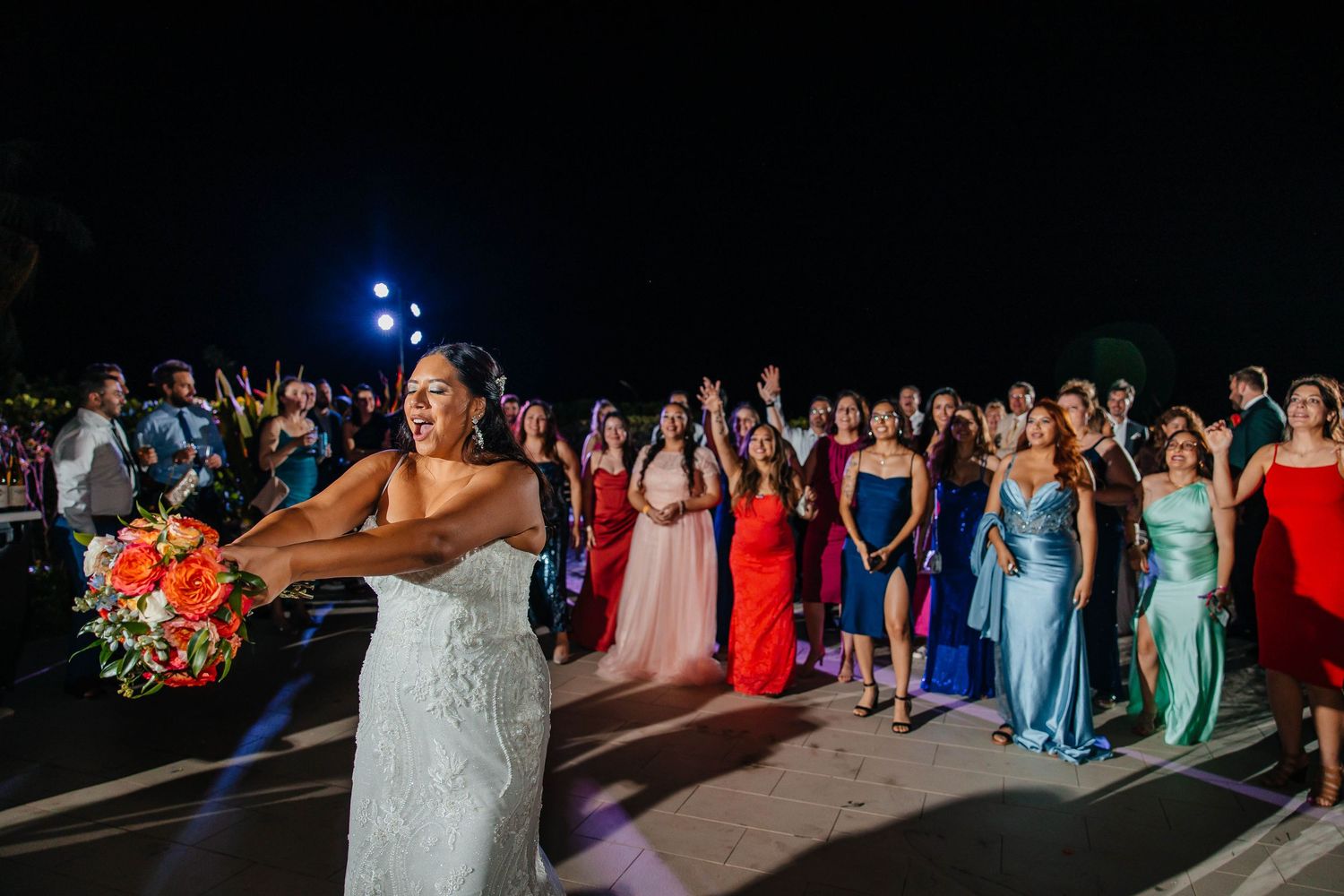 Bride in white lace gown tosses bouquet to guests in colorful dresses at outdoor wedding reception at night.