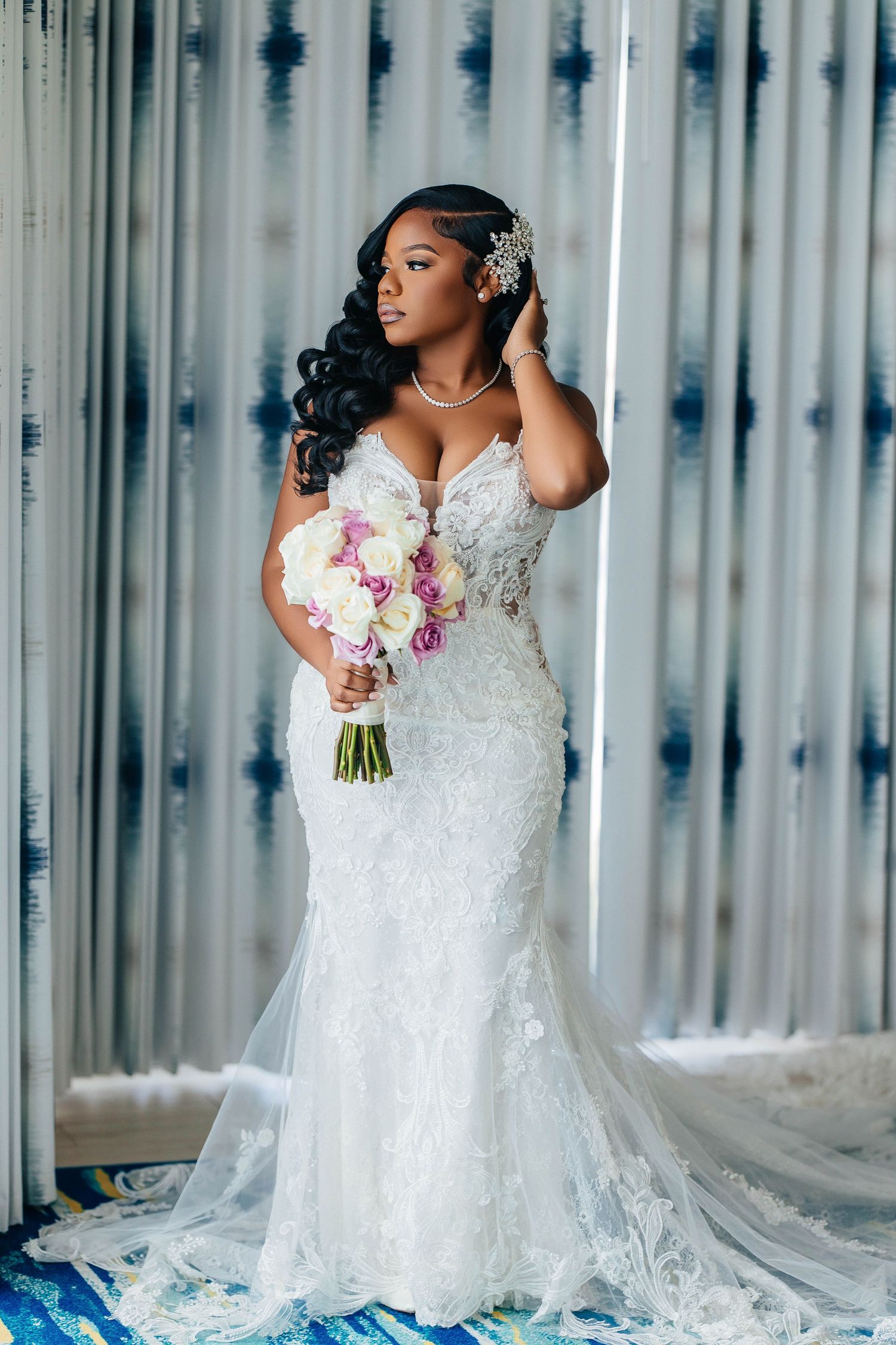 Elegant Black bride in lace mermaid wedding dress holding bouquet, posing by curtains with hair accessory.