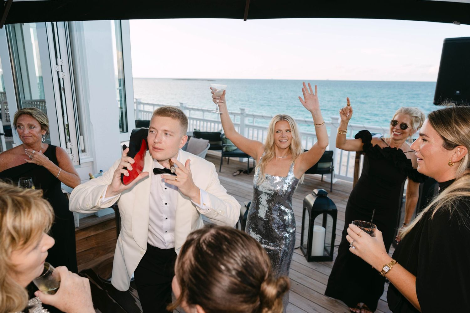 Guests cheer and dance at a seaside party as a man in a white blazer sings into a microphone beside a woman in a sparkly dress.