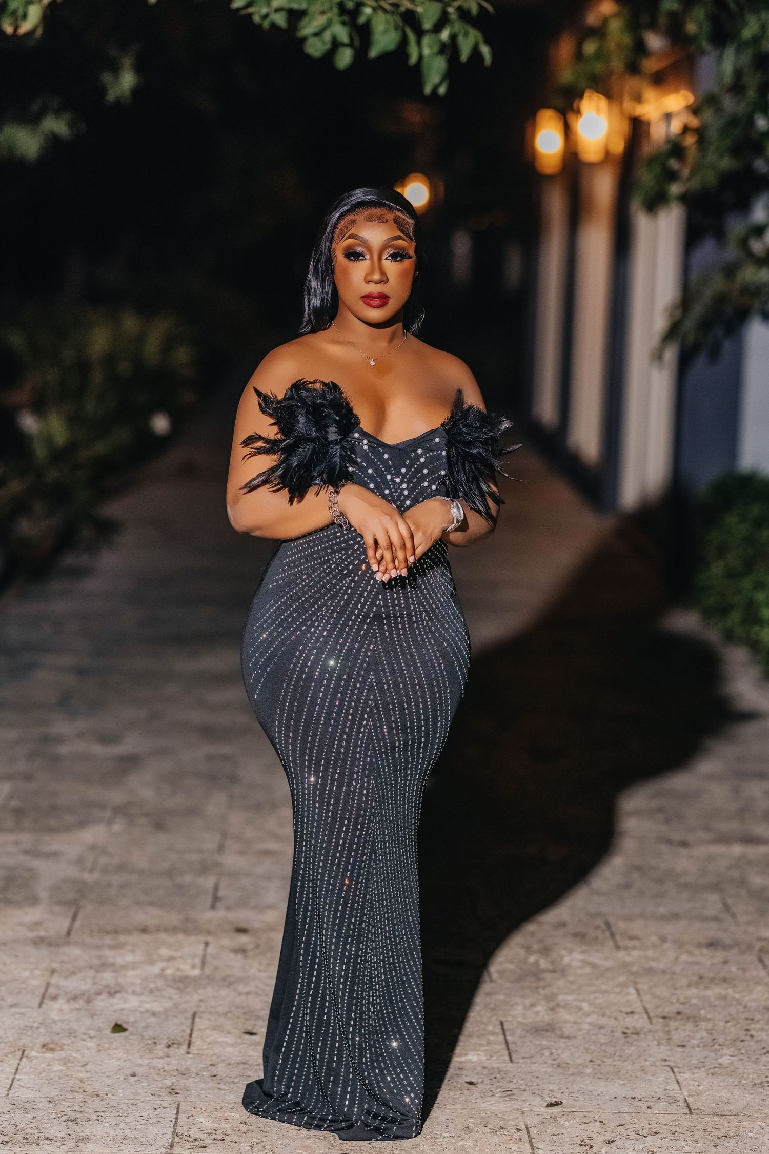 Elegant woman poses in a fitted black sequined gown with feather details and sweetheart neckline at night.