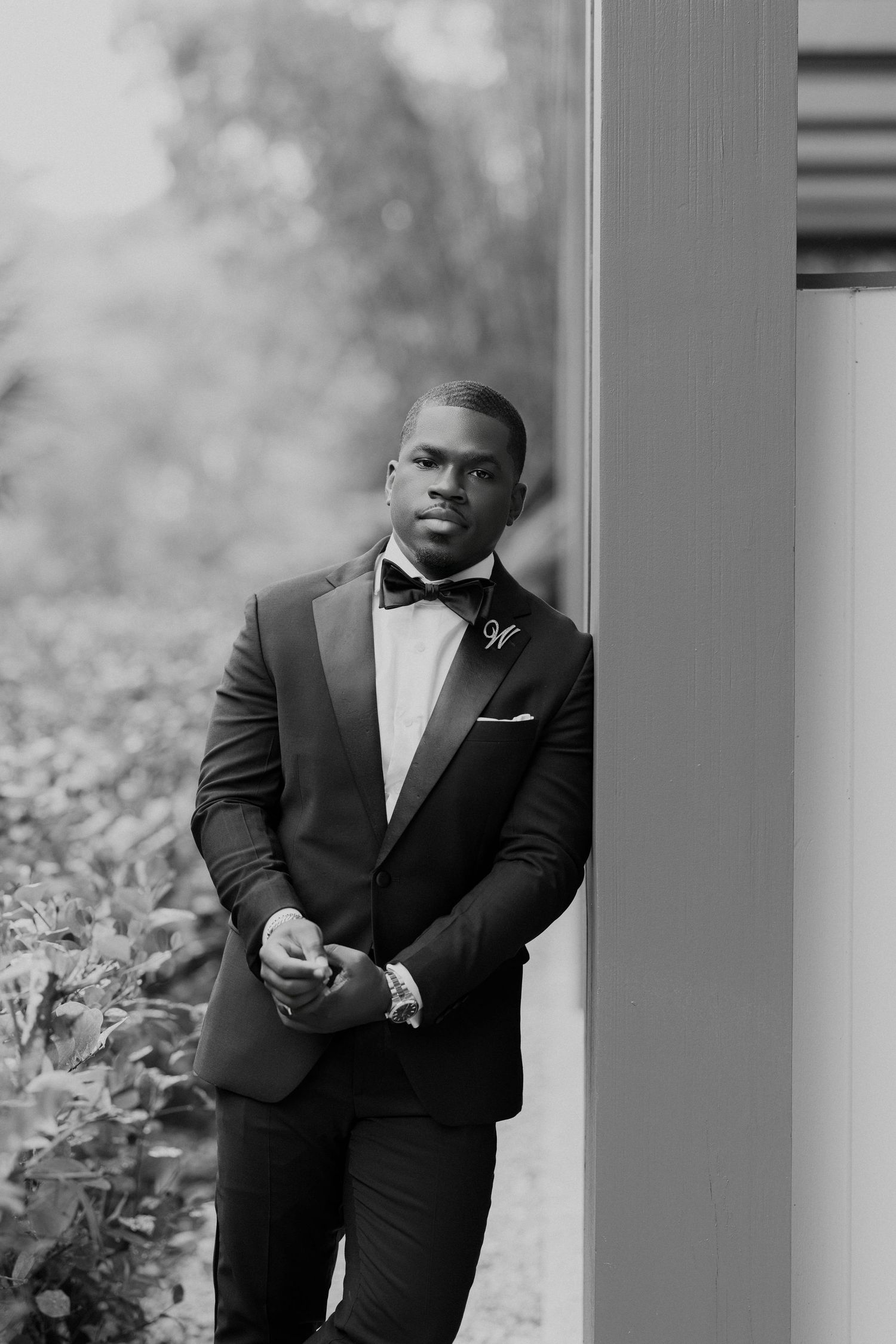Black and white portrait of a well-dressed man in a tuxedo with bow tie and boutonniere leaning against a pillar.