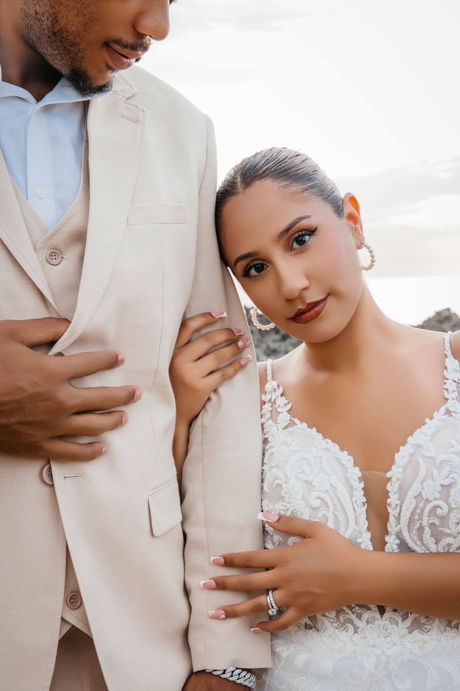 Close-up portrait of newlyweds showing off wedding attire and rings against coastal backdrop.