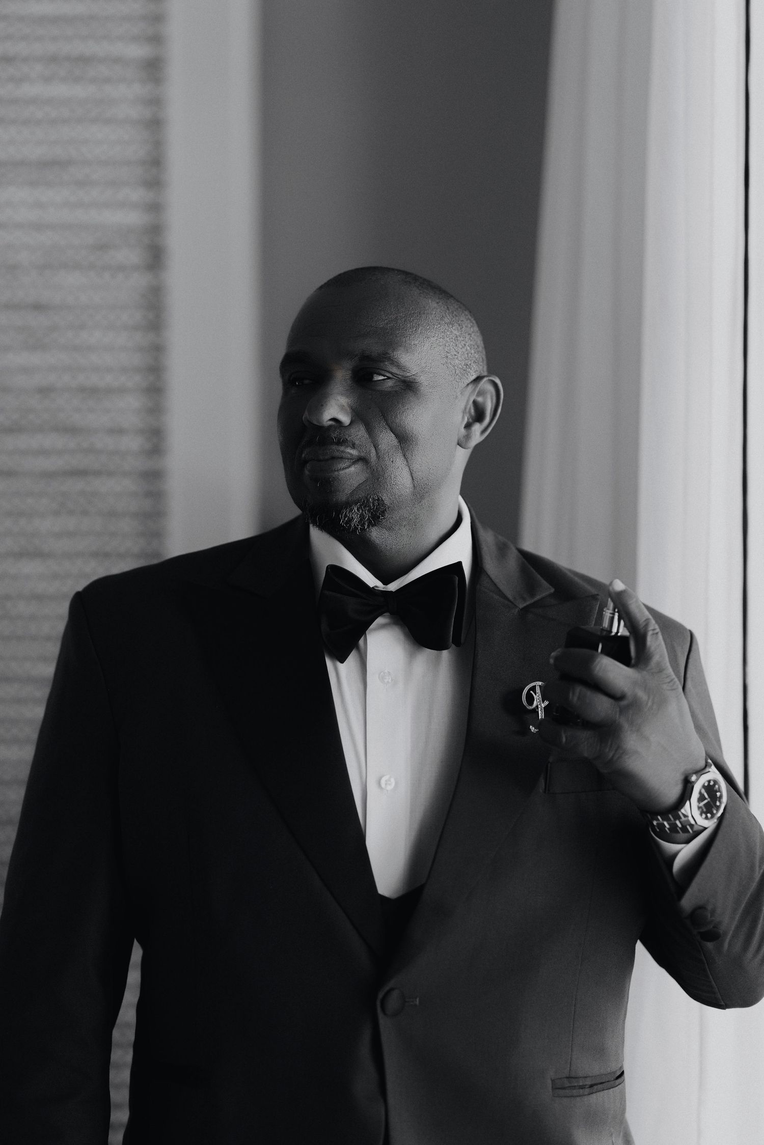 Black and white portrait of a bald man in a tuxedo with bow tie, holding a drink and wearing a luxury watch.
